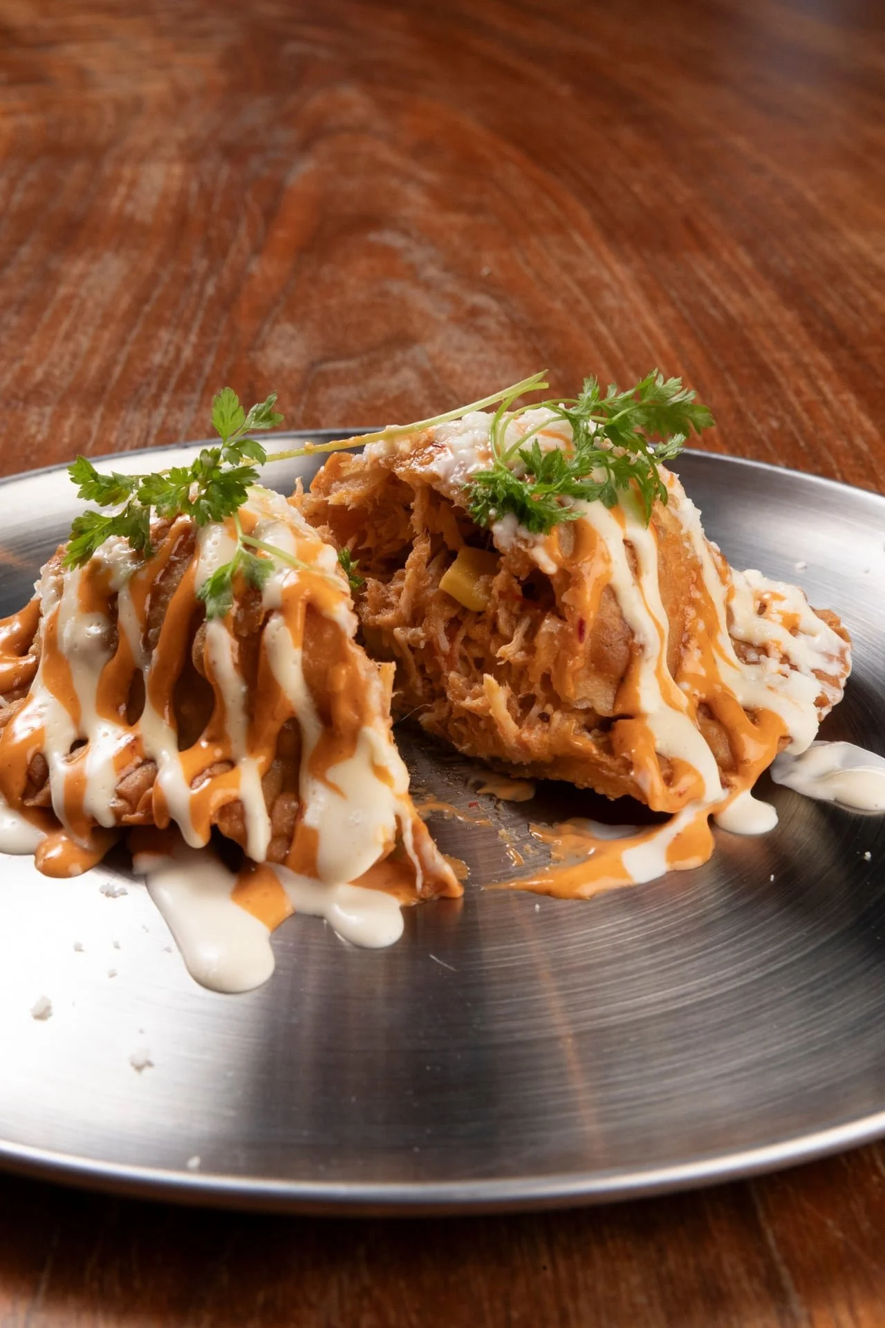 Close-up of two halves of a tamale with shredded meat filling, topped with white and orange sauces, garnished with cilantro, on a metal plate.