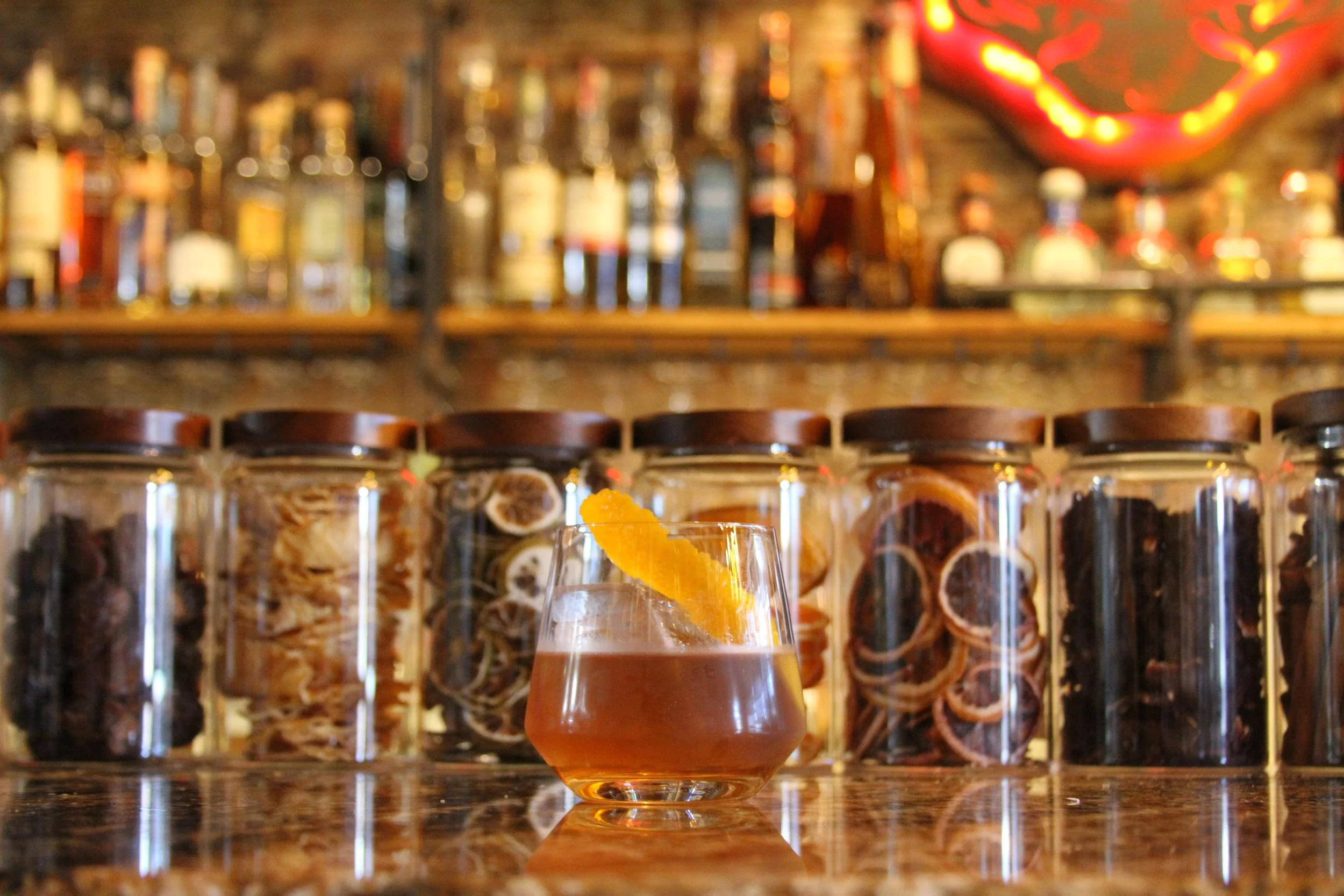 A glass of orange-colored drink with an orange twist garnish in front of a bar with jars containing various ingredients or spices, and blurred bottles on shelves in the background.