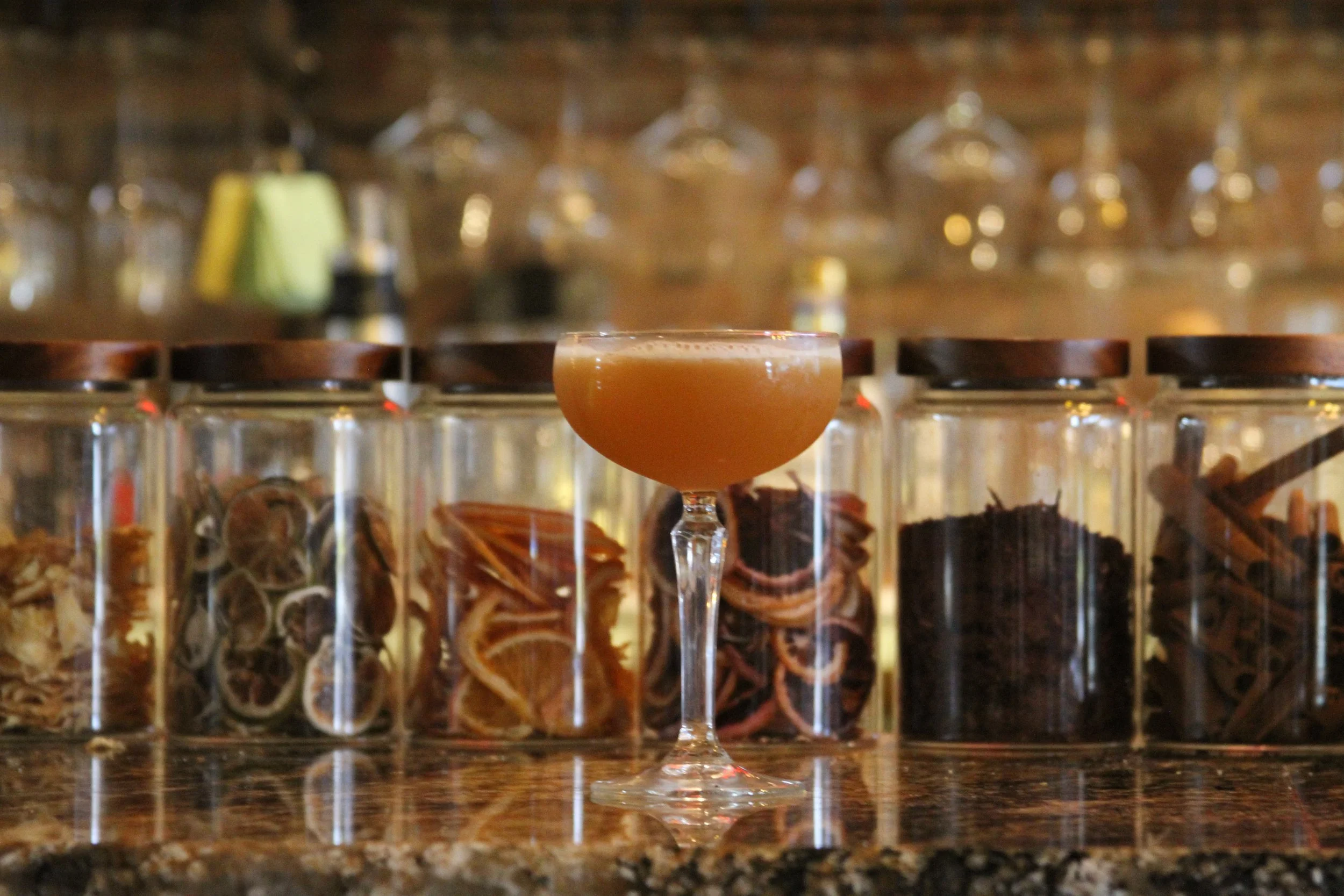 A cocktail in a coupe glass with an orange hue, placed on a bar counter. Behind it, there are glass jars filled with dried fruit slices and other bar ingredients, with a blurred background of glasses and barware.