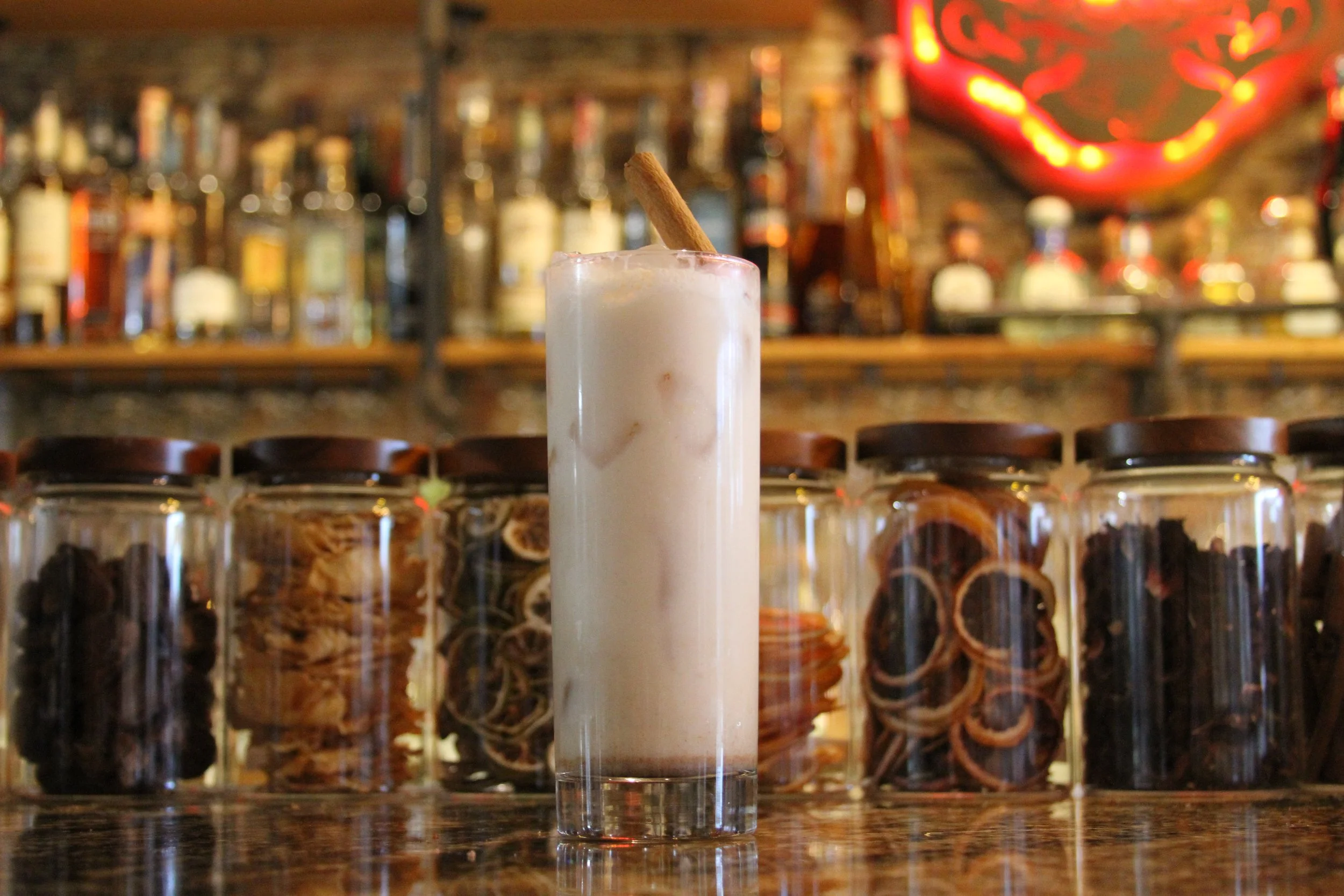 A tall glass of a creamy, frothy beverage with a cinnamon stick garnish, set on a bar counter with blurry bottles and neon sign in the background.