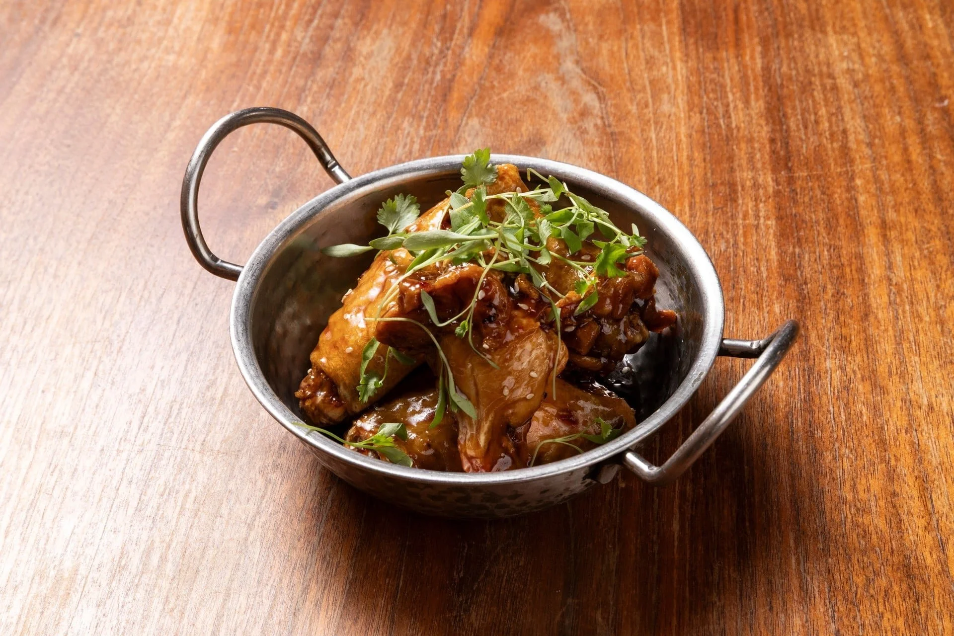 A small metal bowl with handles containing a cooked dish topped with fresh herbs, placed on a wooden table.