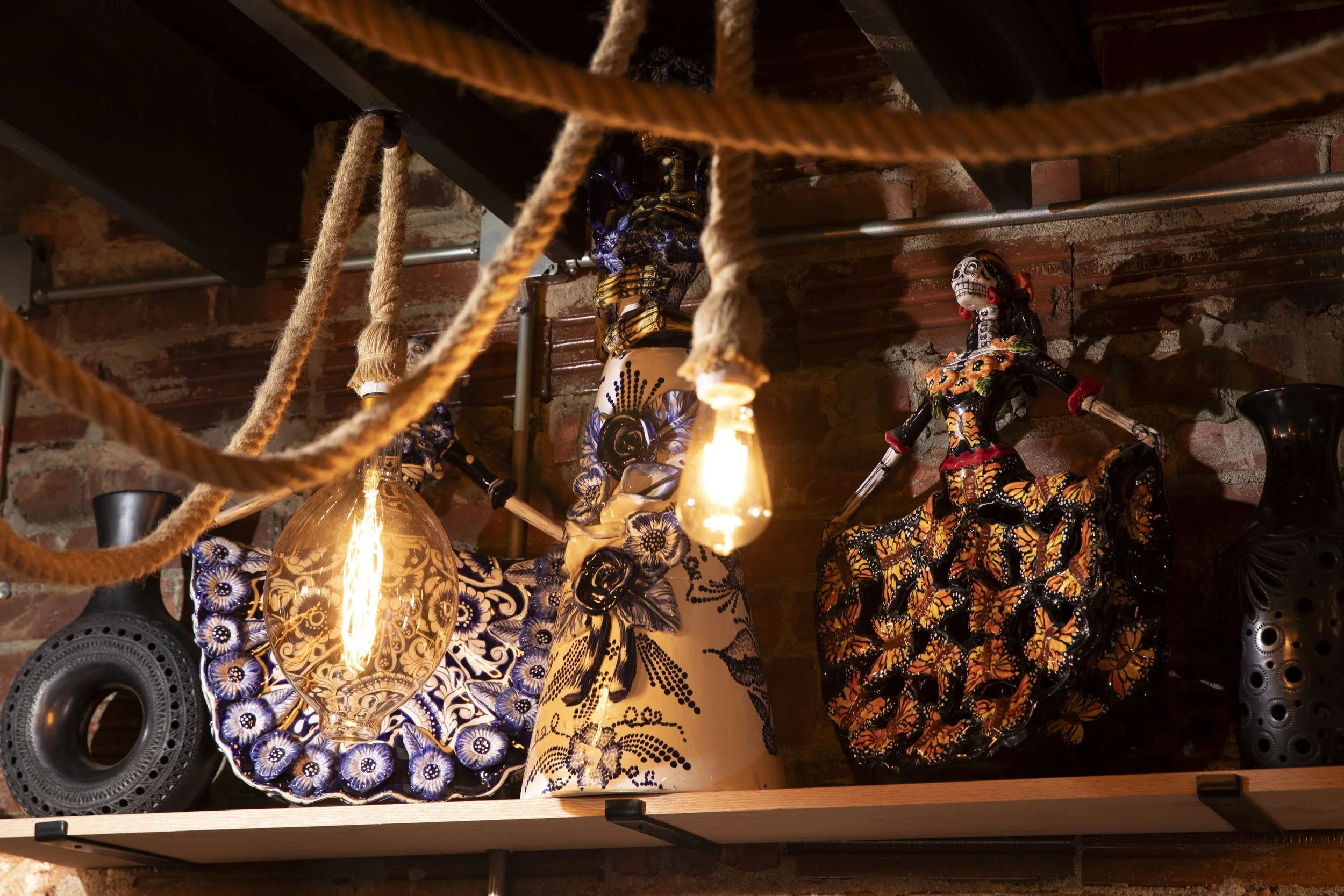 Decorative ceramic figurines of women in traditional Mexican attire, with floral and butterfly patterns, on a wooden surface. Surrounding objects include a glass lamp, black plates, and a brick wall background with hanging ropes and speakers.