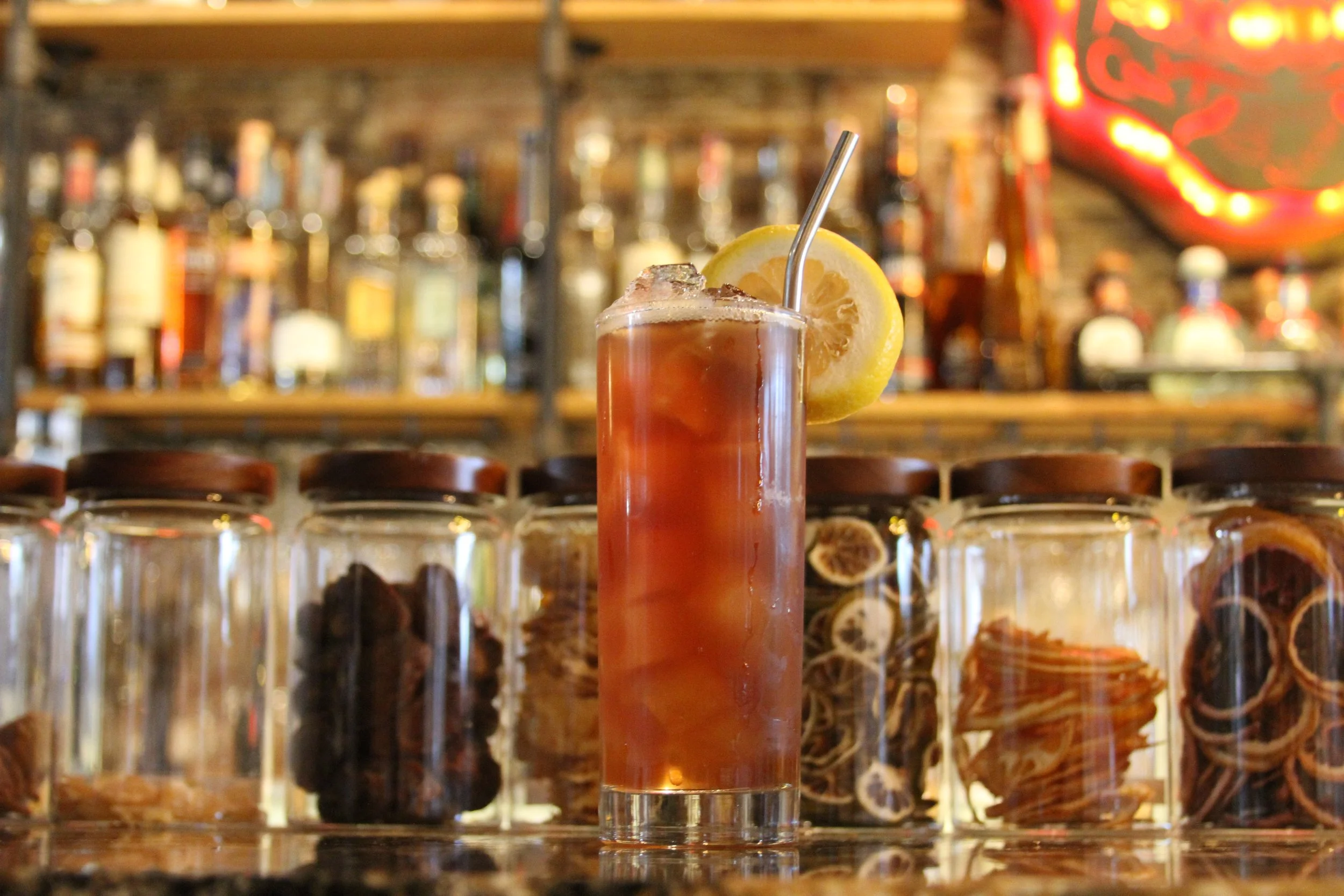 A tall glass of iced tea with a lemon slice and metal straw on a bar counter, with bottles of liquor blurred in the background.