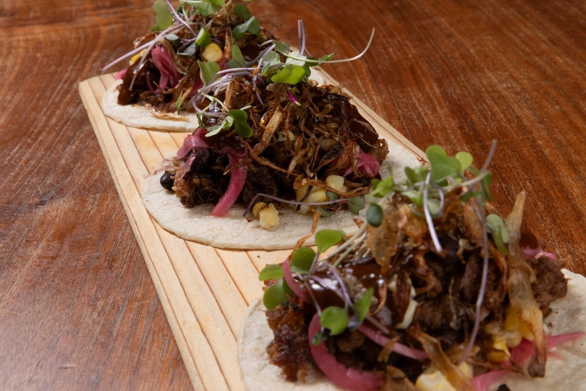 Three small tacos on a wooden serving board topped with shredded roasted meat, microgreens, and thinly sliced pickled onions, on a wooden table.