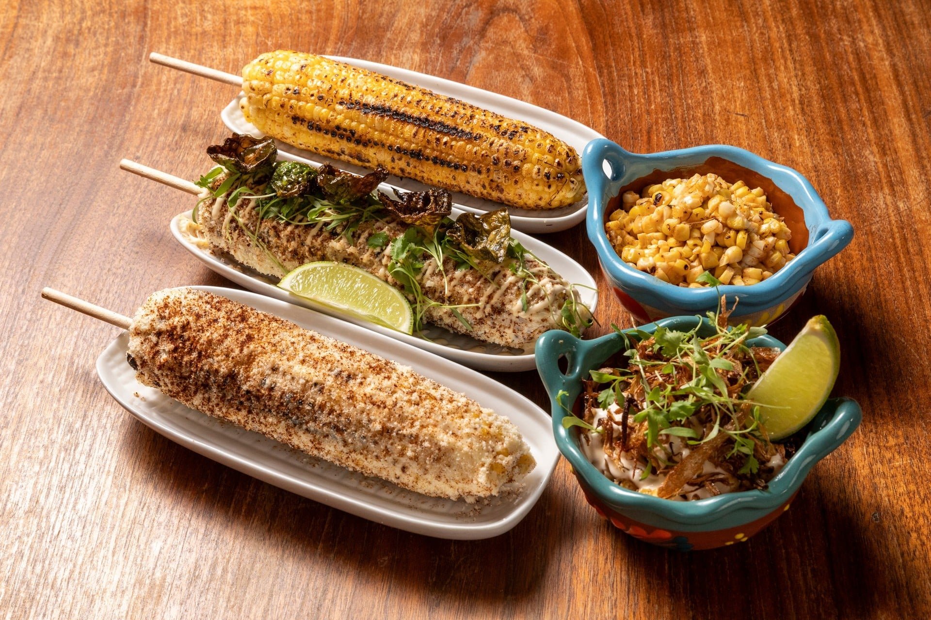 Corn on the cob, grilled, served with lime wedge, in white dishes and colorful bowls, on a wooden table.
