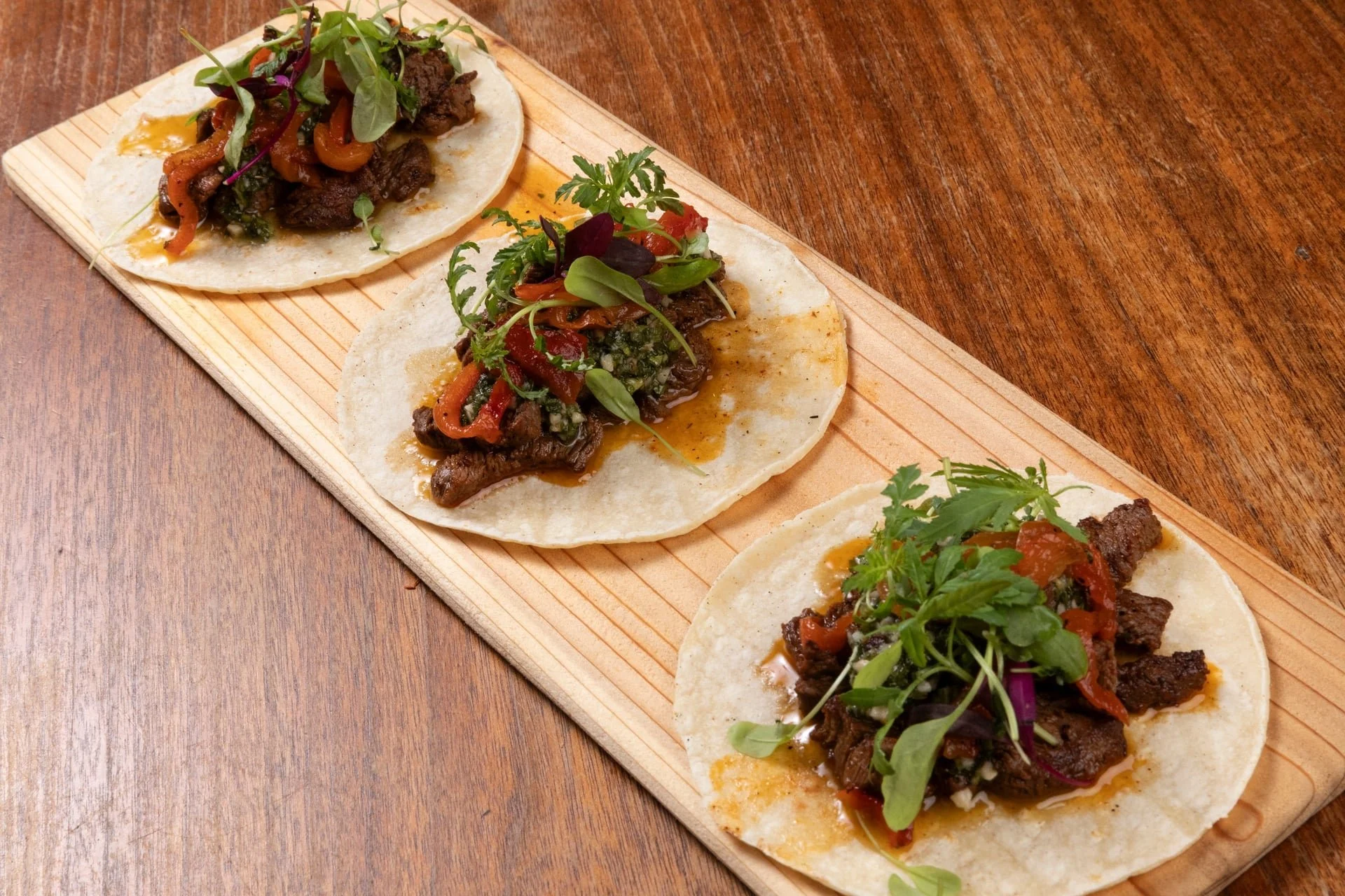 Three beef tacos topped with chopped cilantro, microgreens, and sautéed peppers on a wooden serving tray.