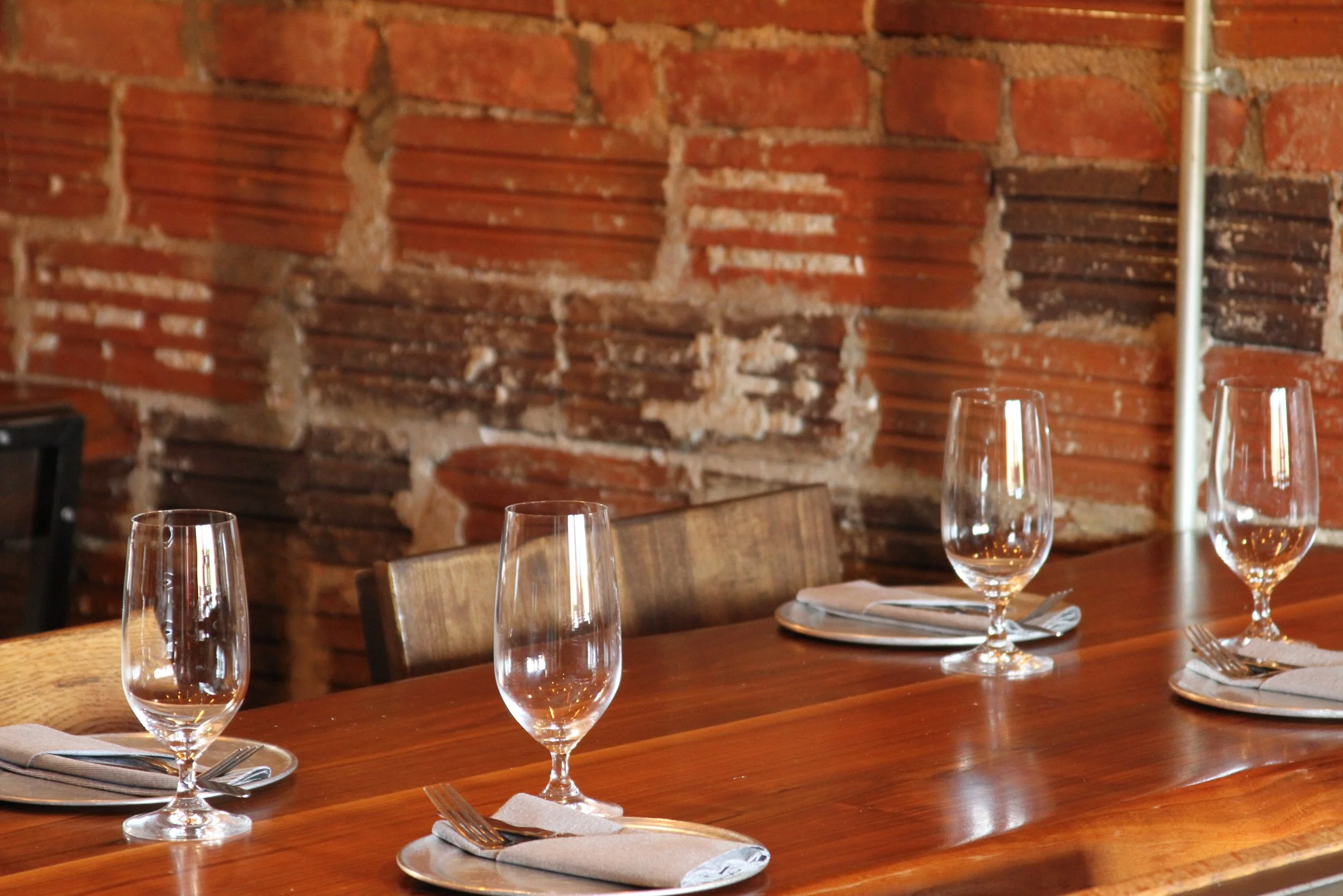 A dining table set with empty wine glasses, napkins, and silverware in front of a brick wall with exposed brick detailing.