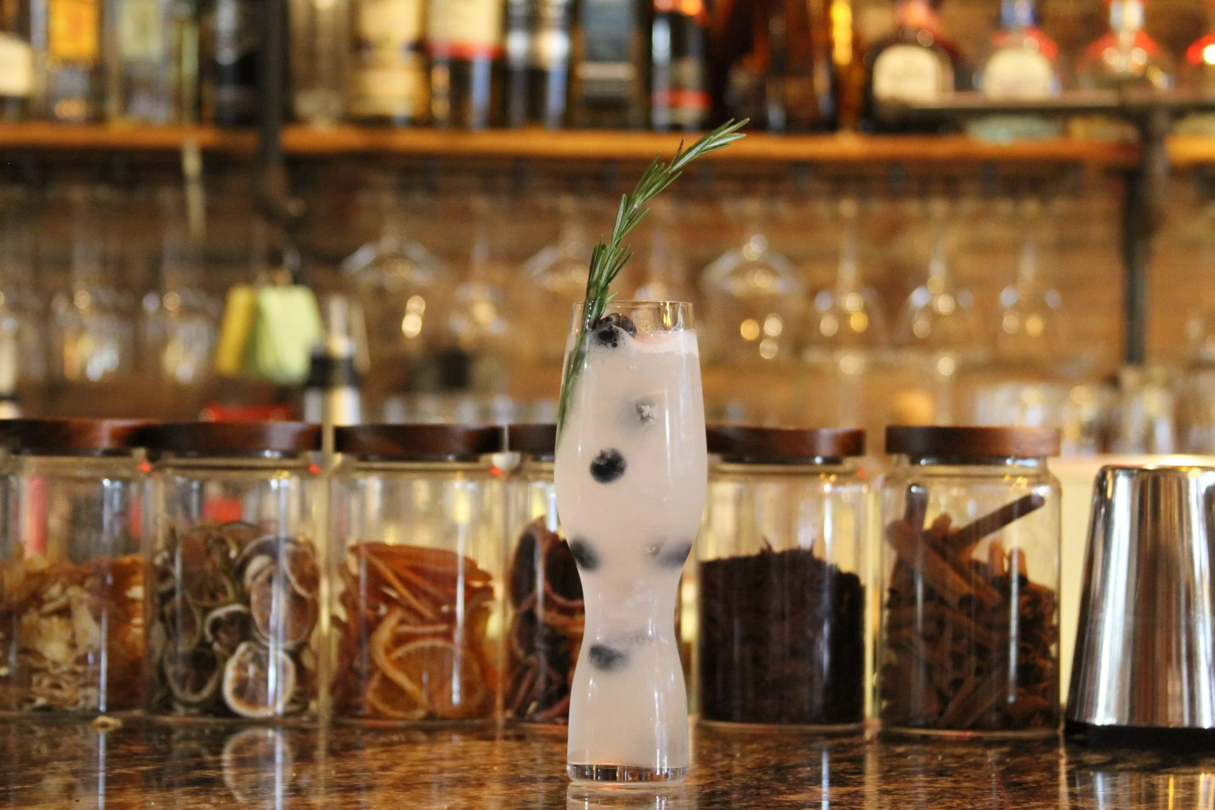 A tall glass of blueberry and herbal cocktail garnished with a sprig of rosemary, set on a bar counter with a blurred background of bottles and barware.