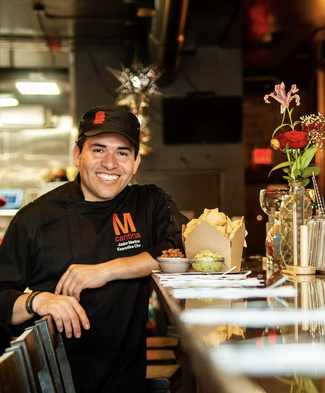 Smiling chef in black uniform and cap behind a bar, with bowls of chips, guacamole, and salsa on the counter, decorated with a vase of flowers in the restaurant.