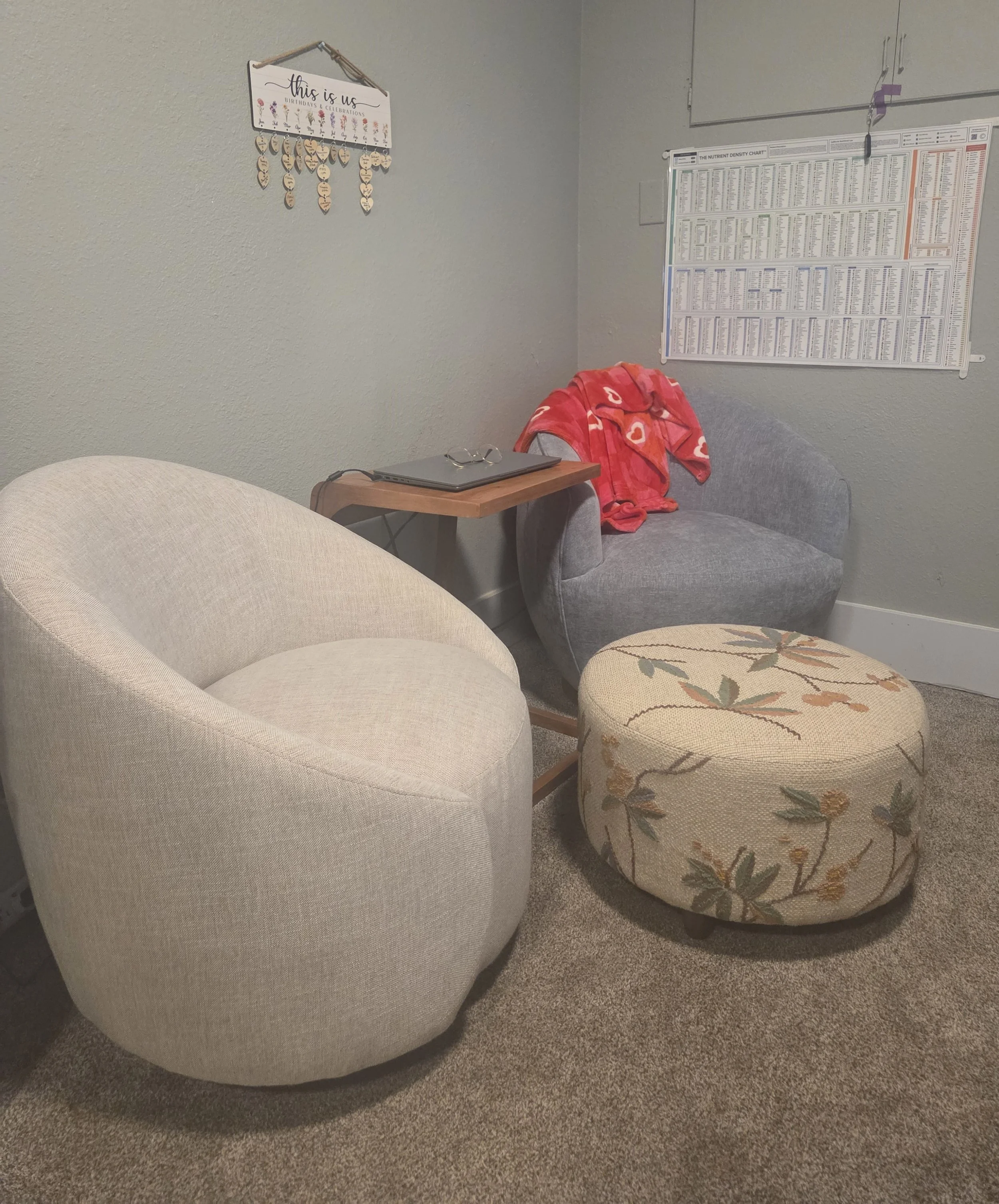 A cozy seating area with two armchairs, a floral ottoman, a small wooden side table with glasses and a laptop, a wall hanging, and a nutrient density chart on the wall.