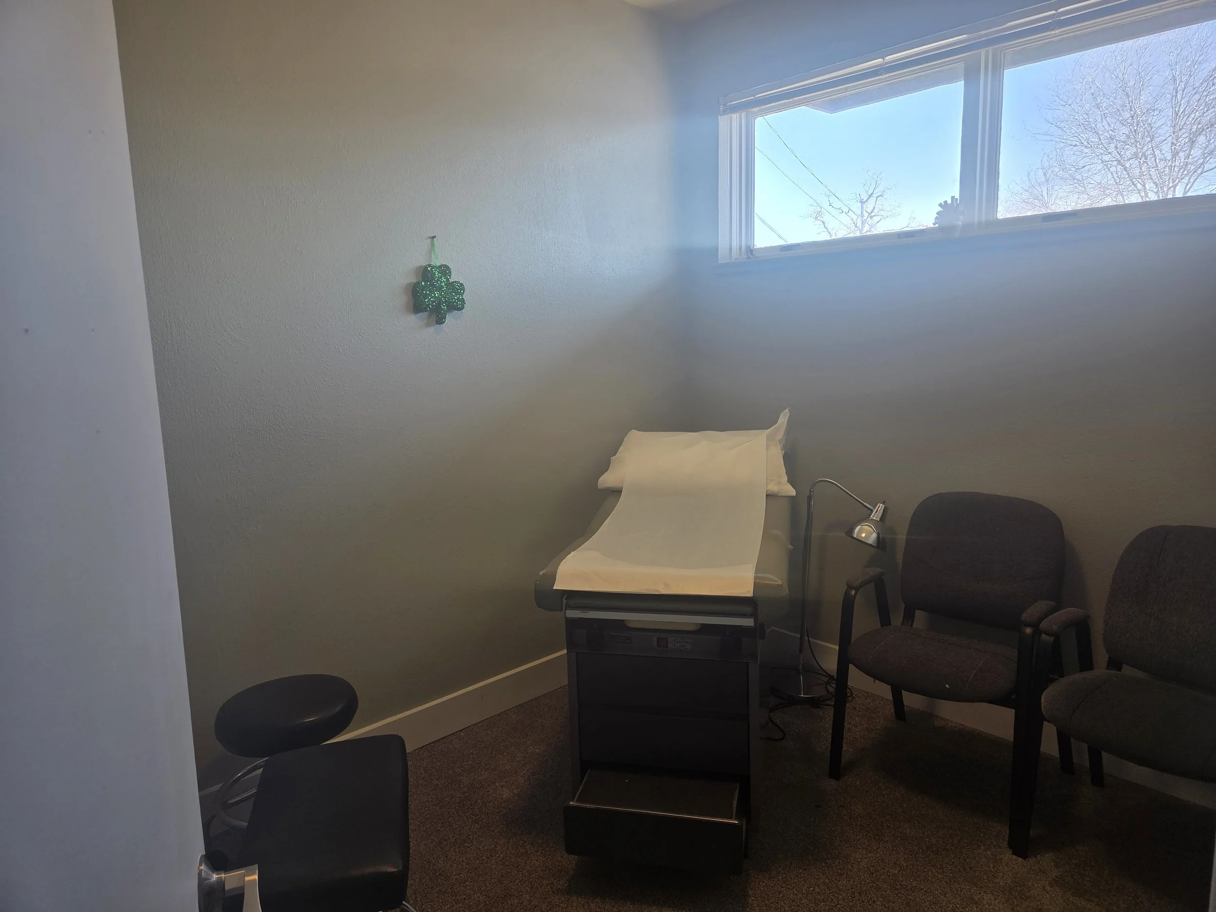 Medical examination room with an examination table covered in paper, two chairs, a stool, a small lamp, and a Craftsman-style decoration on the wall. There is a large window letting in natural light.