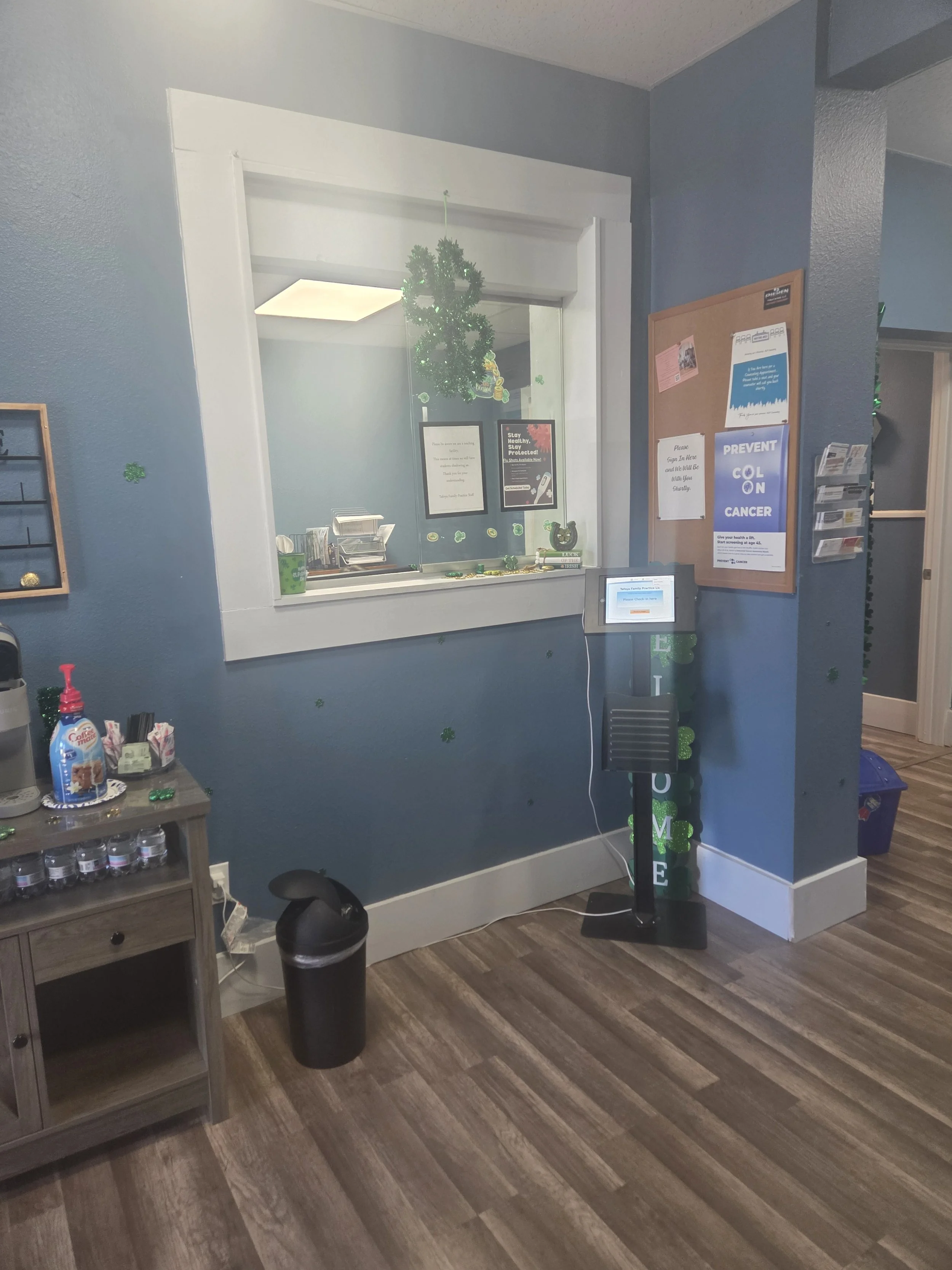 A reception area with a blue wall and a window countertop decorated with a green wreath. There is a small table with condiments and snacks, a trash can, and a digital check-in kiosk with a stand. Various signs and notices are posted on the wall, some