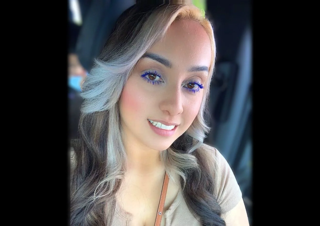 A smiling young woman with fair skin, long wavy hair that is blonde with dark roots, and wearing makeup with blue mascara and eye makeup, sitting inside a vehicle.