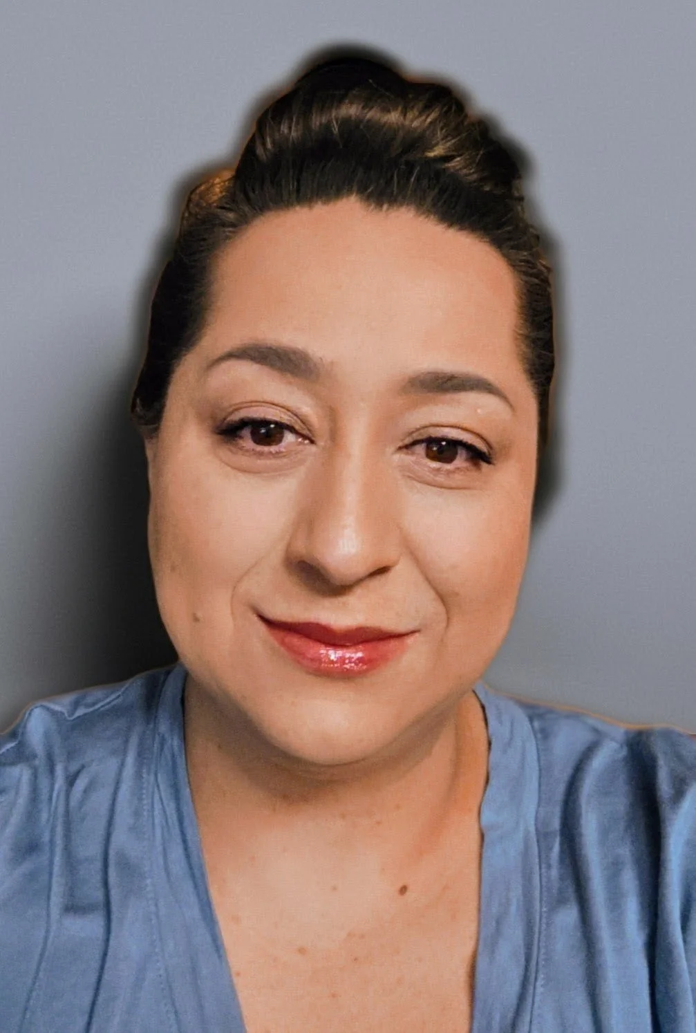 A woman with dark brown hair styled in an updo, wearing makeup and a light blue top, smiling at the camera against a plain, gray background.