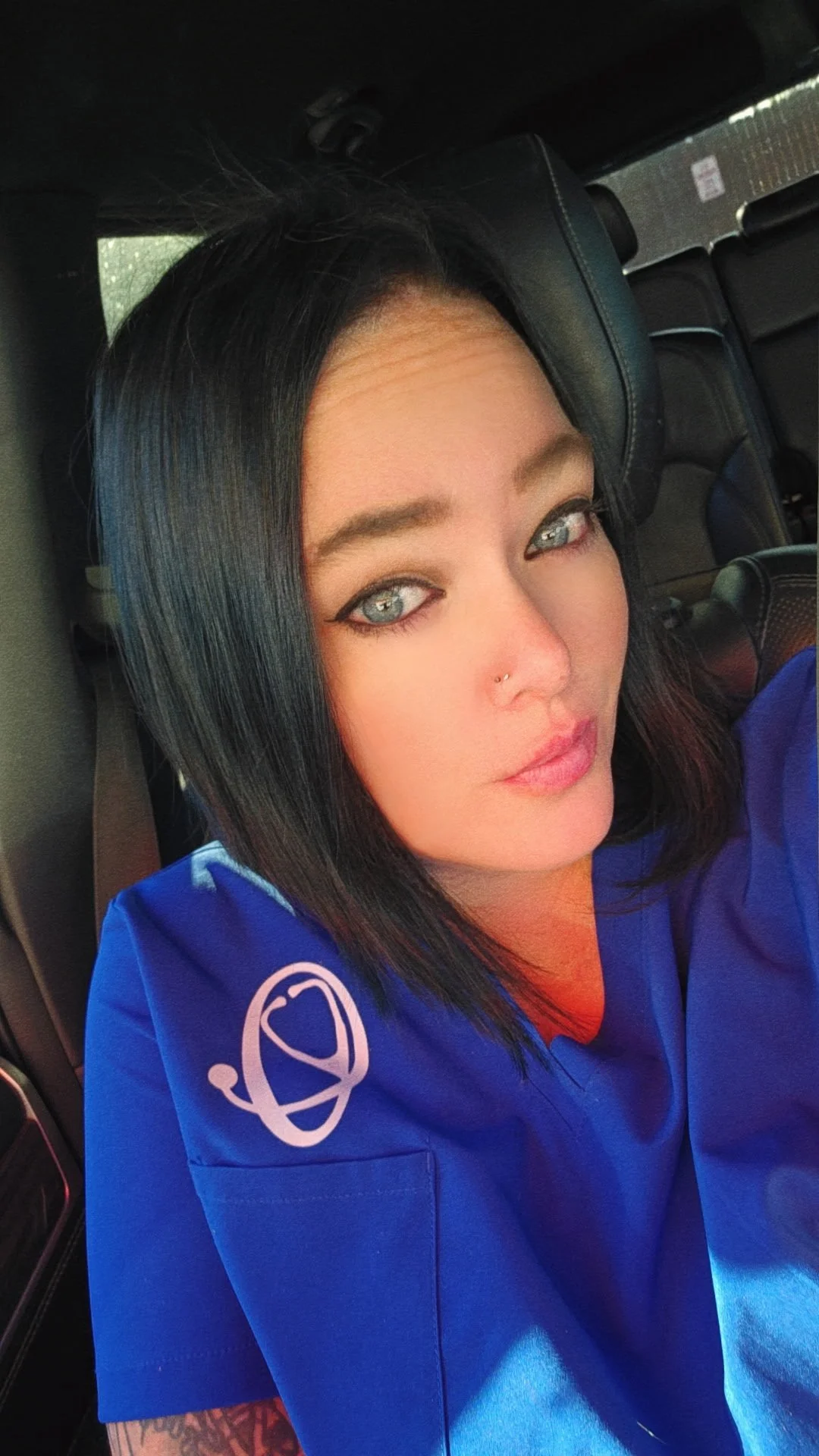 A woman with dark hair and blue eyes sitting in a vehicle, wearing a blue uniform with a logo on the sleeve.