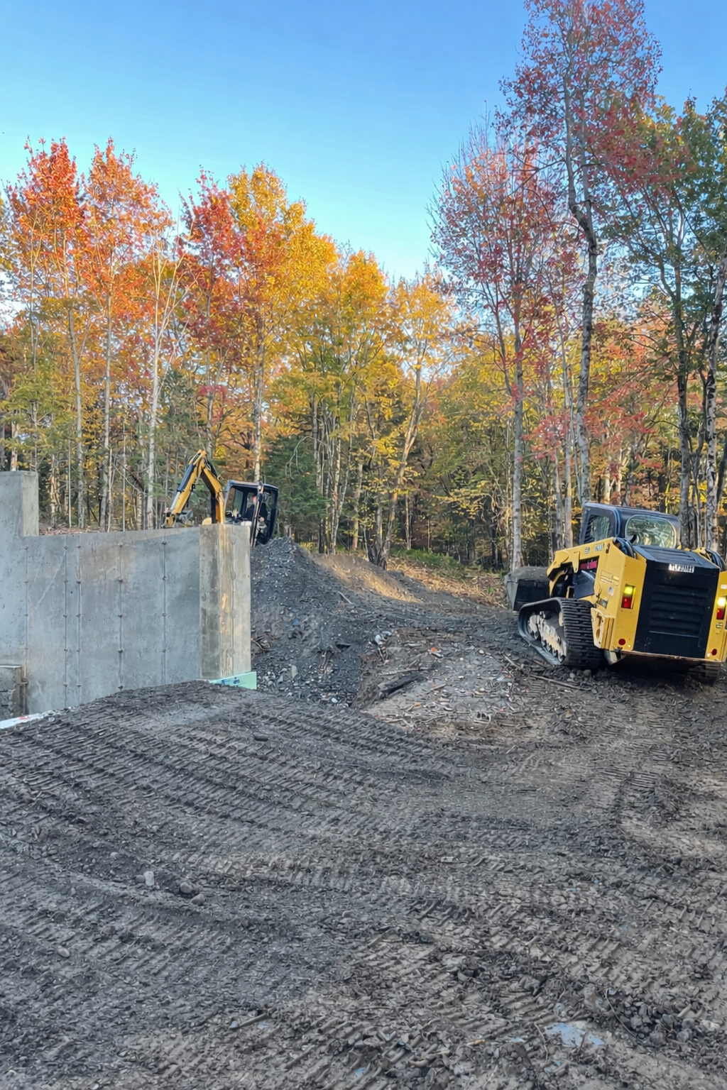 Construction site in a forested area featuring a small yellow bulldozer and an excavator, with autumn trees in the background.