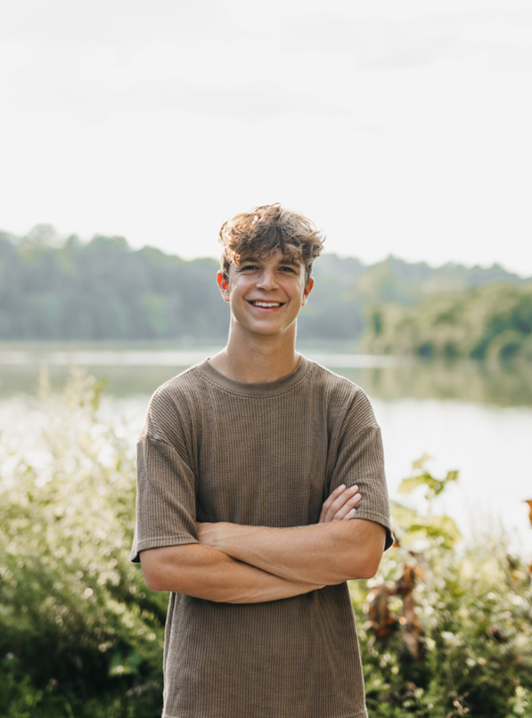 A young man with curly hair standing outdoors near a body of water, smiling and crossing his arms.