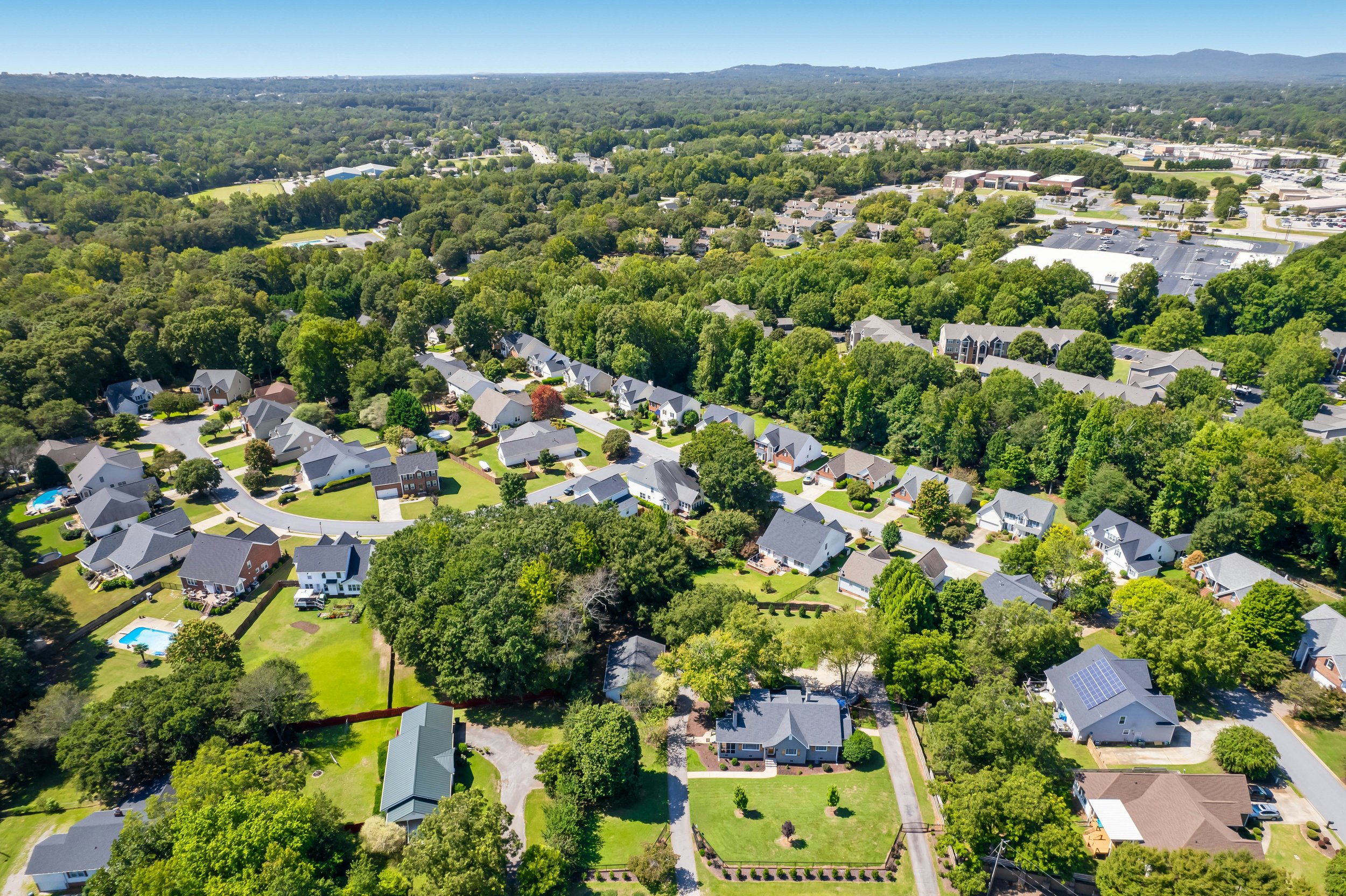 Aerial view of a suburban neighborhood with houses, green lawns, and trees, with a forested area behind and a commercial area in the distance.