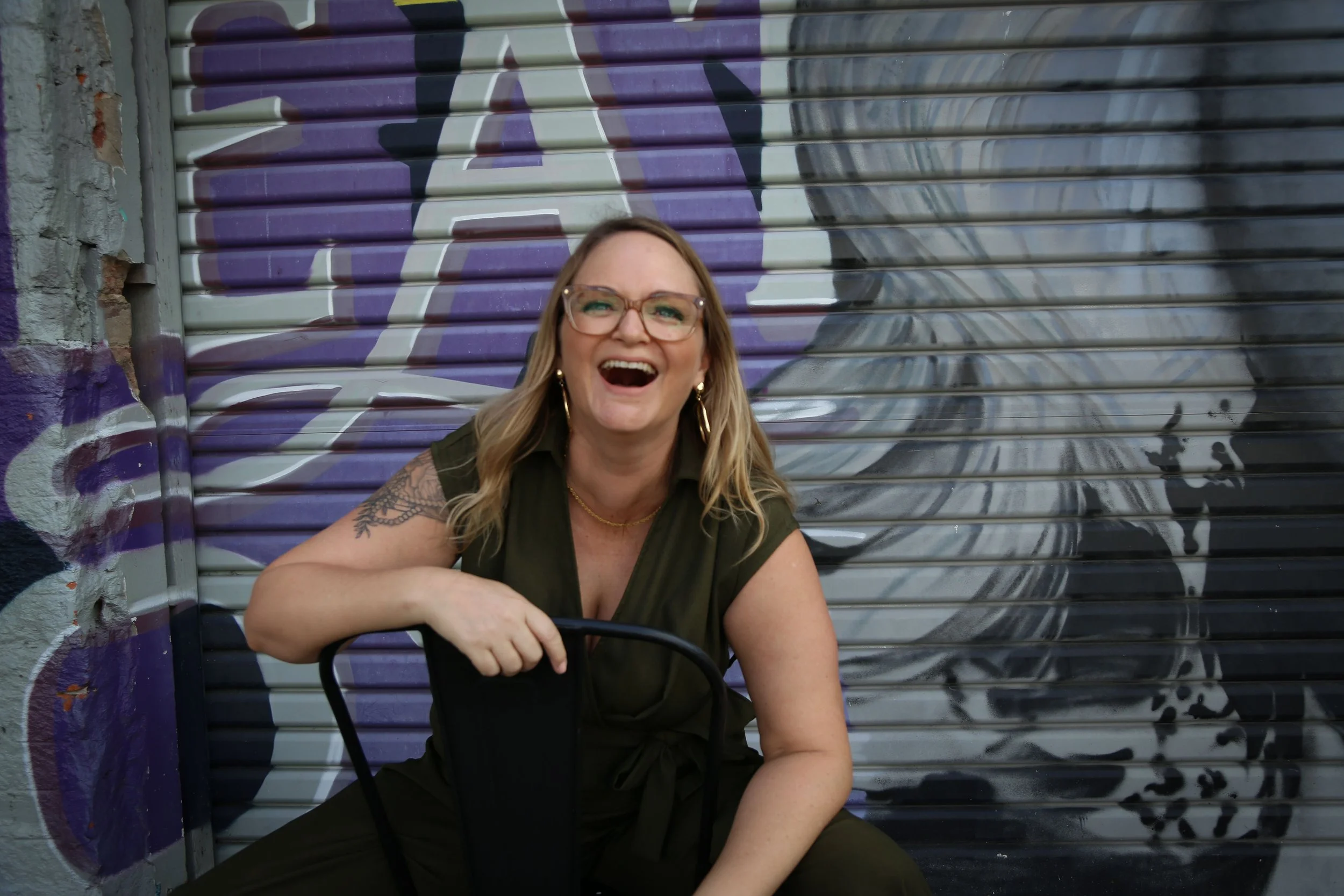 Woman with glasses and earrings laughing, seated with a black chair in front of a purple and black graffiti mural on a metal rolling door.