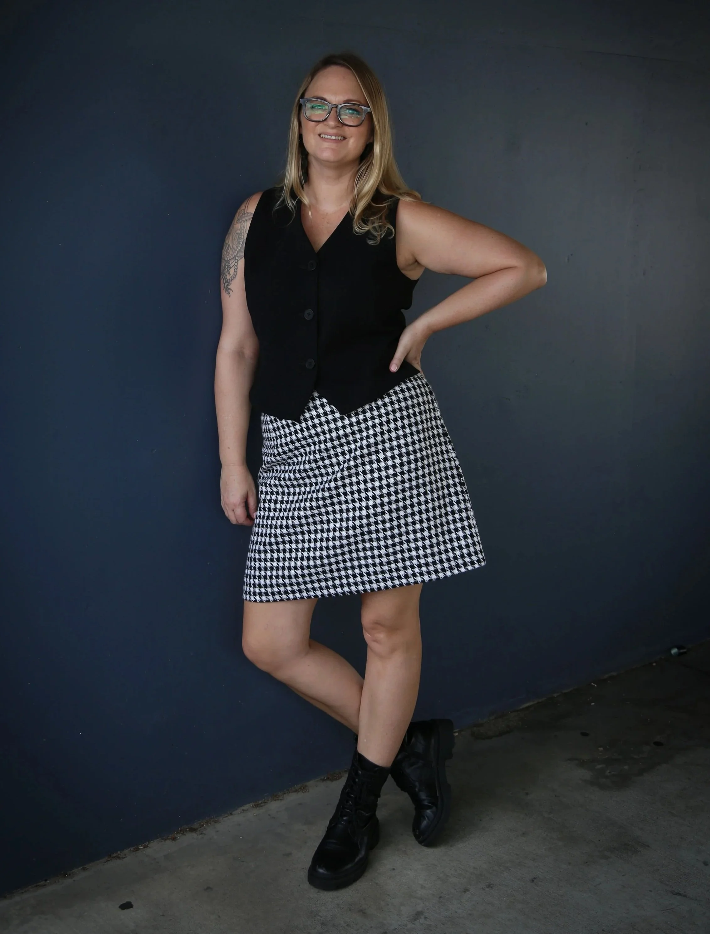 Woman standing against a dark wall, wearing glasses, a sleeveless black vest, a black and white checkered skirt, and black boots, smiling with one hand on her hip.