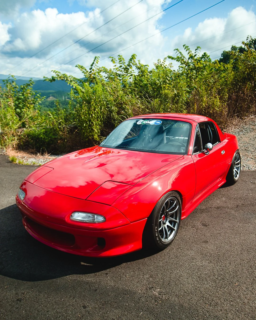Red sports car parked on a paved surface with green bushes and mountains in the background under a partly cloudy sky.