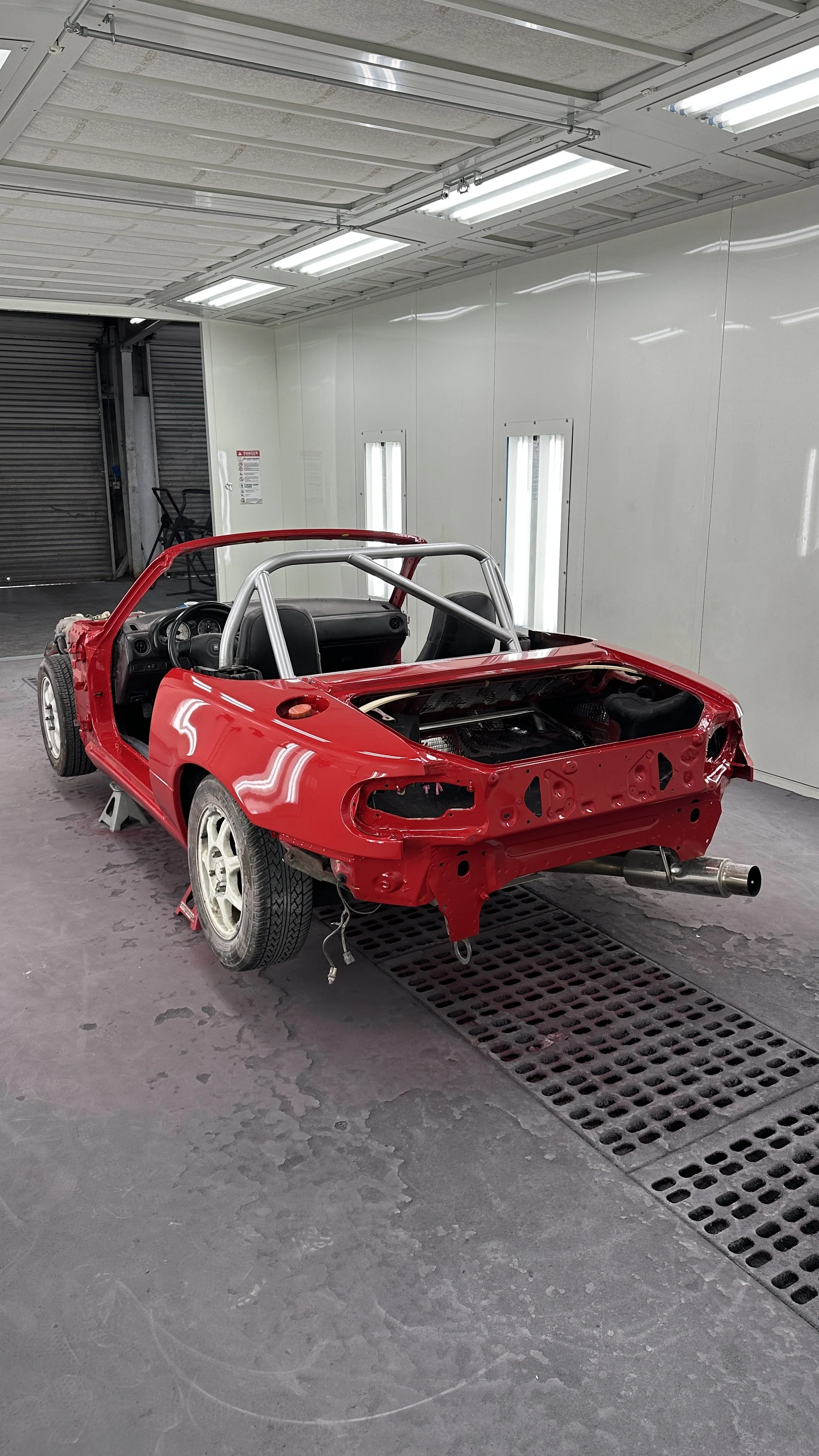 Red race car body shell in a workshop, missing parts and exposed internal frame, on a gray floor with drainage grate, illuminated by bright overhead lights.