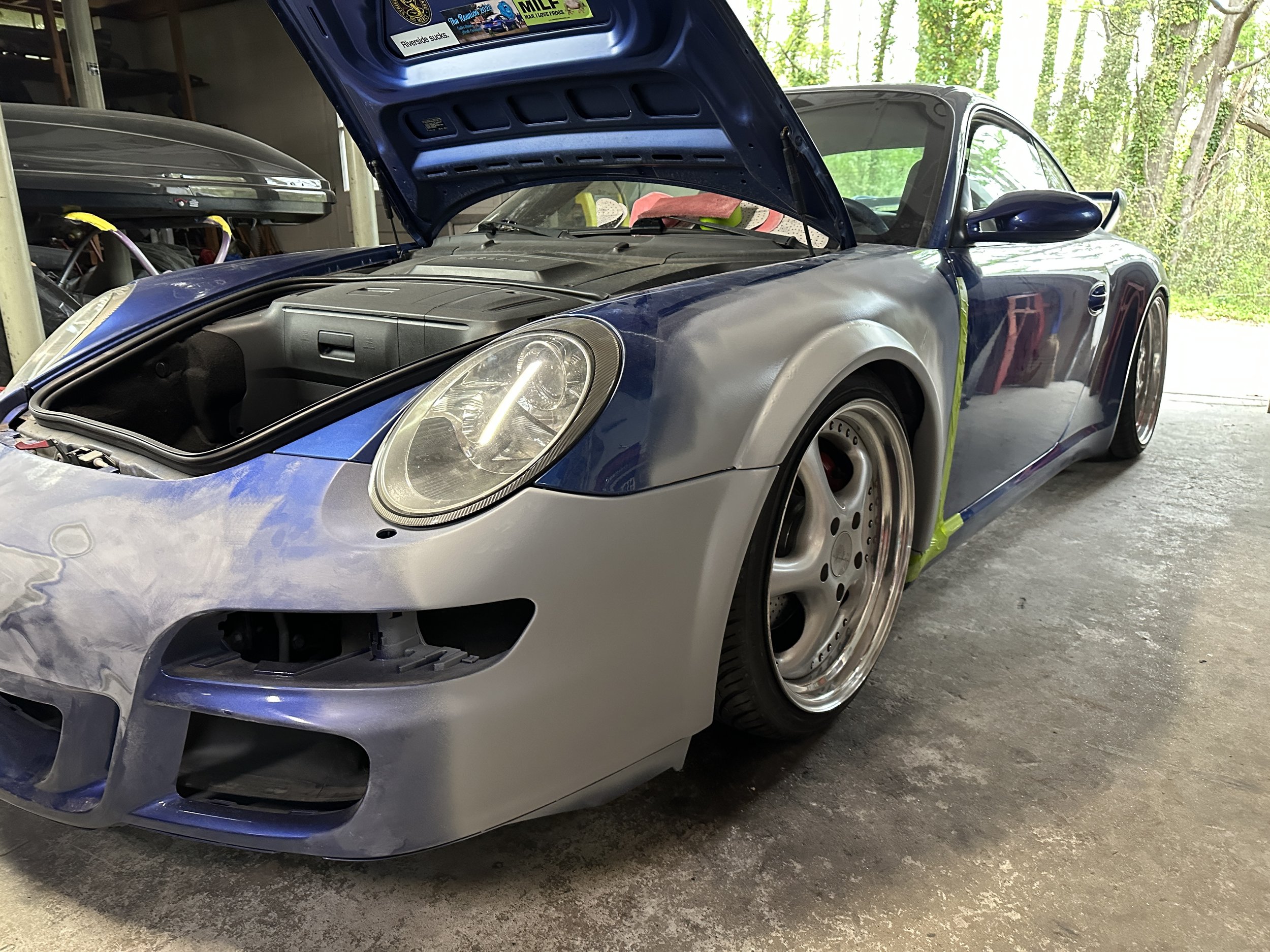 A blue sports car with its hood open, parked inside a garage or workshop space. The car has custom wheels and a sleek design, and there are some tools and storage shelves visible in the background.