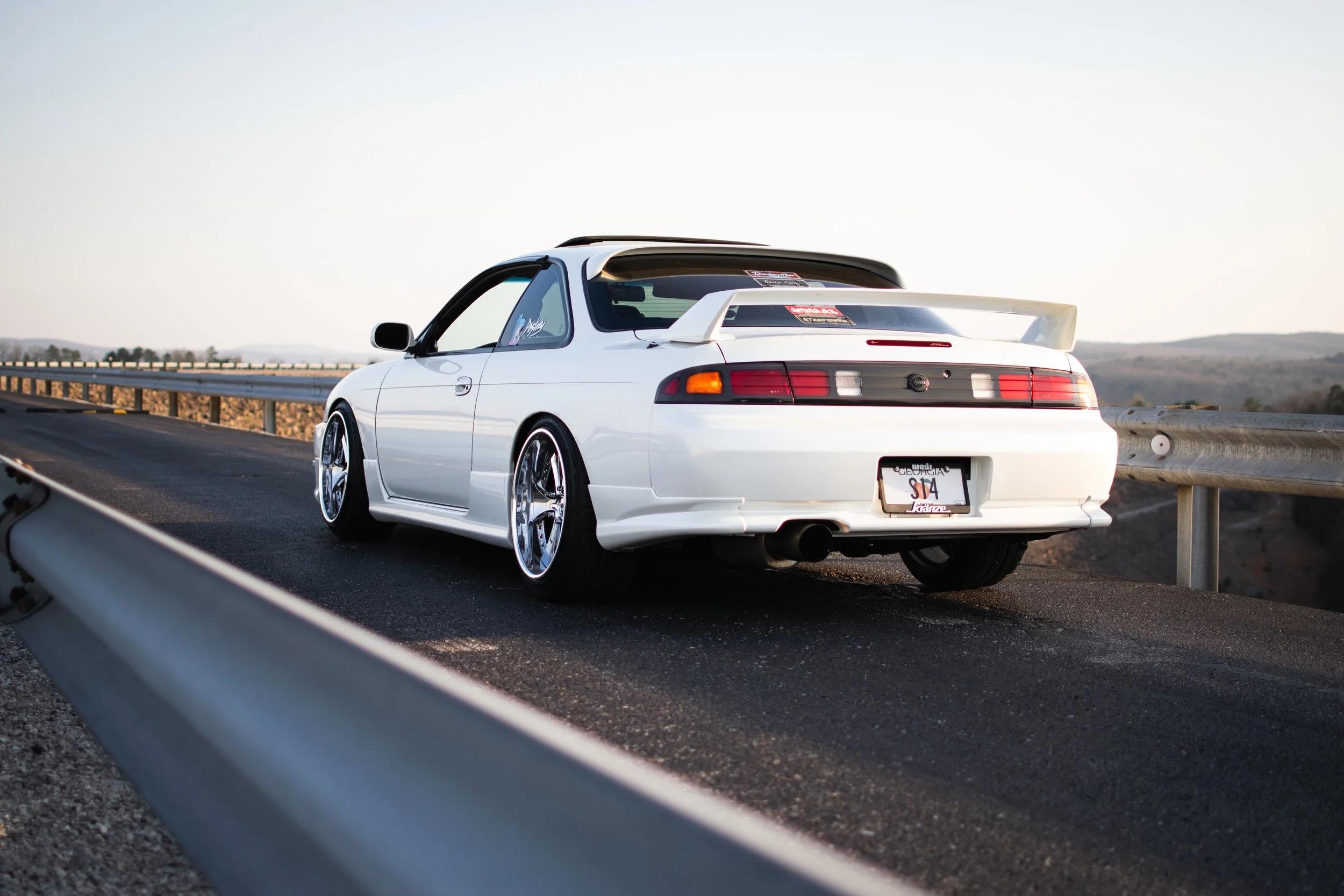 White sports car with a spoiler parked on the side of a highway at sunset.