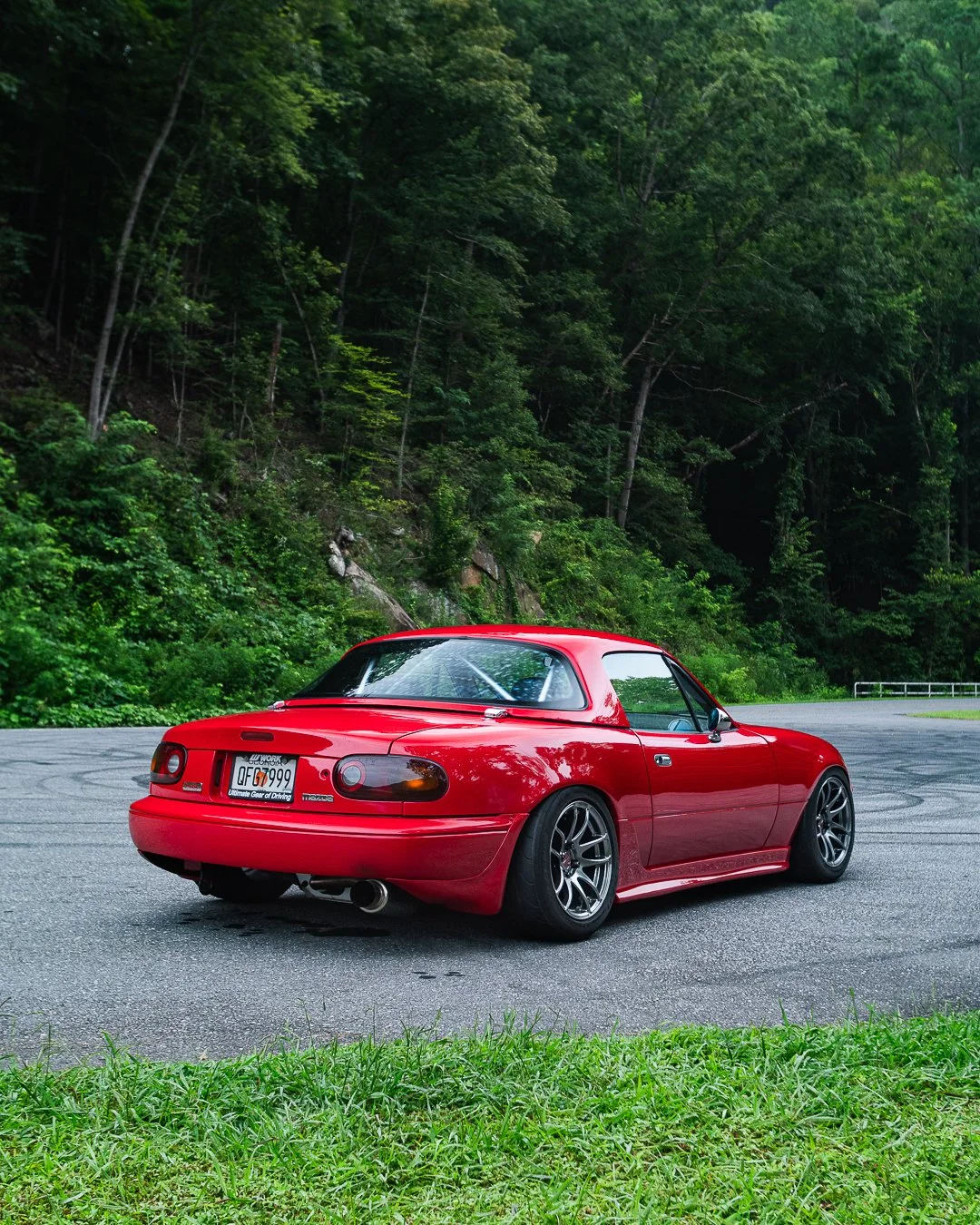 Red vintage Mazda MX-5 Miata sports car parked on a winding road with a lush green forest in the background.