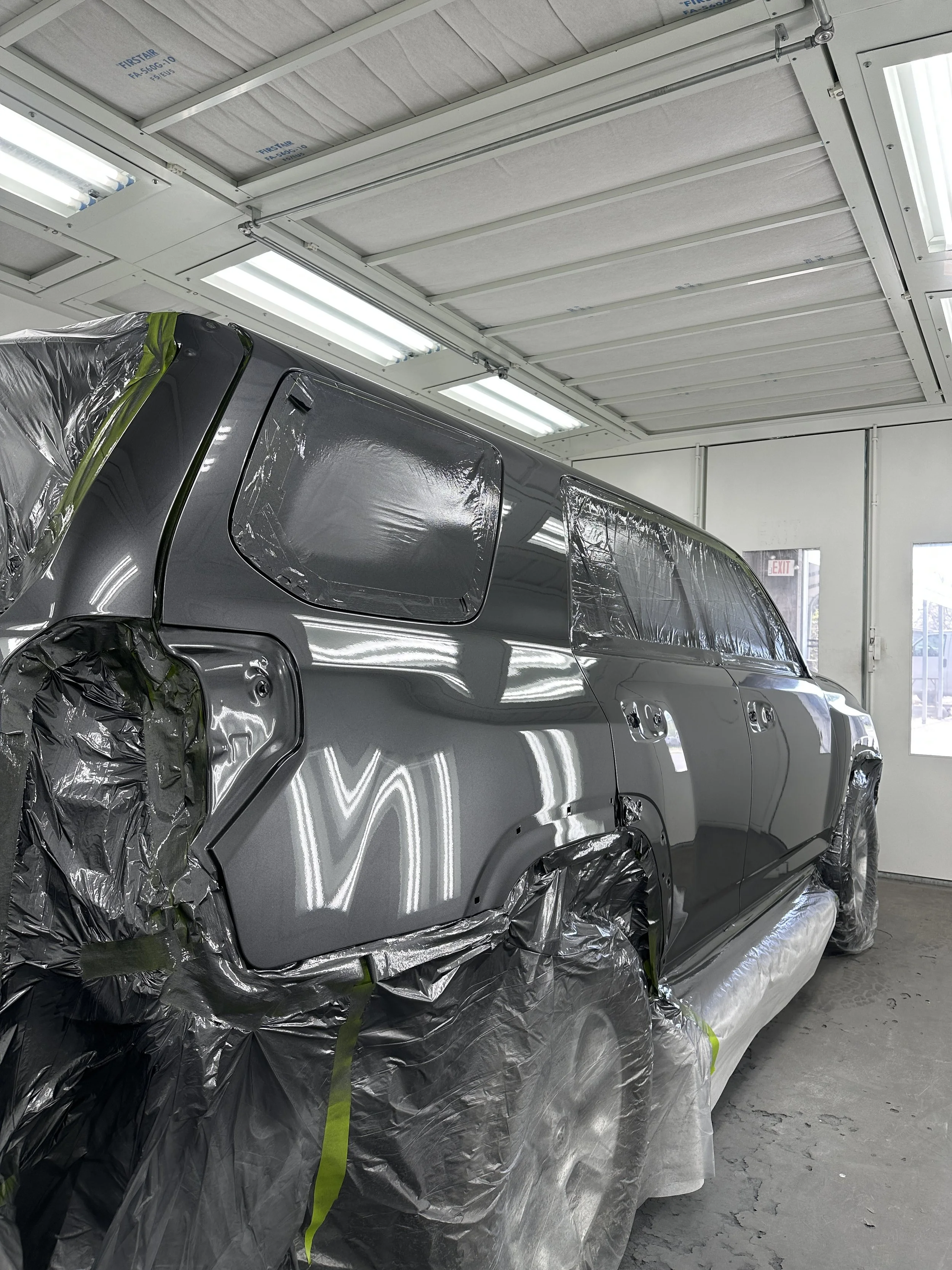 A gray vehicle in a paint booth covered with plastic and masking tape, with a freshly painted glossy finish. The environment is well-lit with fluorescent lights on the ceiling.