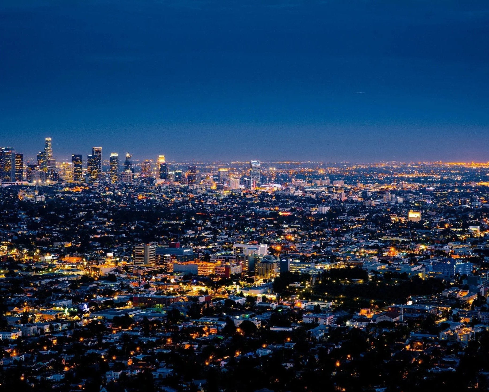 Nighttime aerial view of downtown Los Angeles with illuminated skyscrapers and city lights stretching to the horizon.