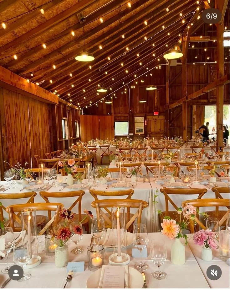 Une salle de réception en bois décorée pour un événement, avec des tables longues, chandelles, vases avec des fleurs, guirlandes lumineuses suspendues au plafond en bois.