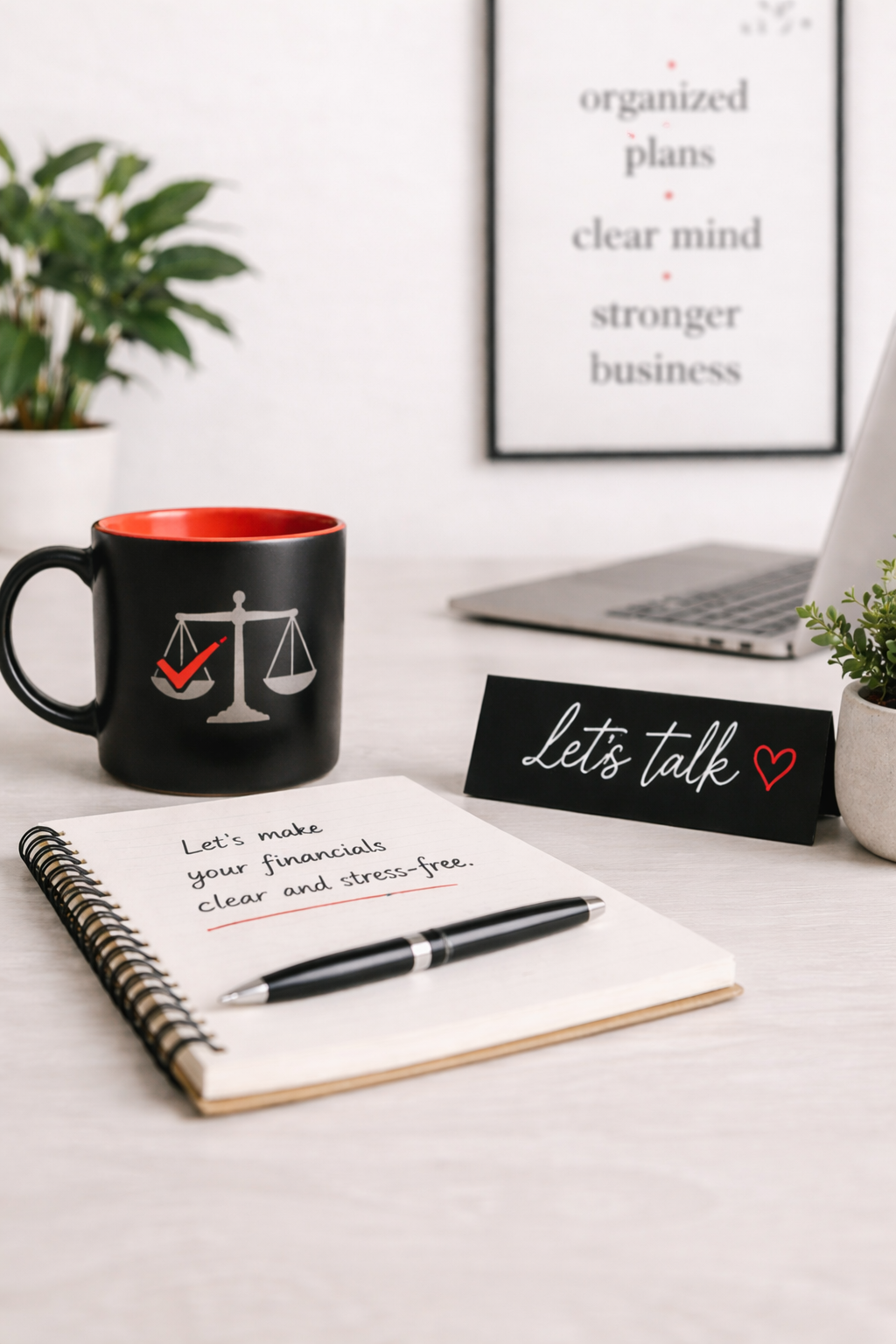 A workspace featuring a black mug with a scale and checkmark design, a notebook with financial planning notes, a pen, a sign with 'Let's talk' and a heart, a framed sign with 'organized plans, clear mind, stronger business', a laptop, and a small potted plant.