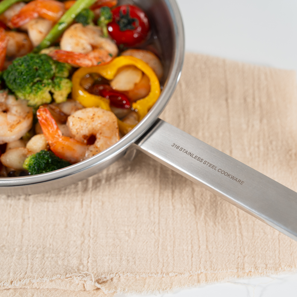 Stainless steel cookware filled with shrimp, broccoli, cherry tomatoes, and yellow bell peppers placed on a beige cloth.