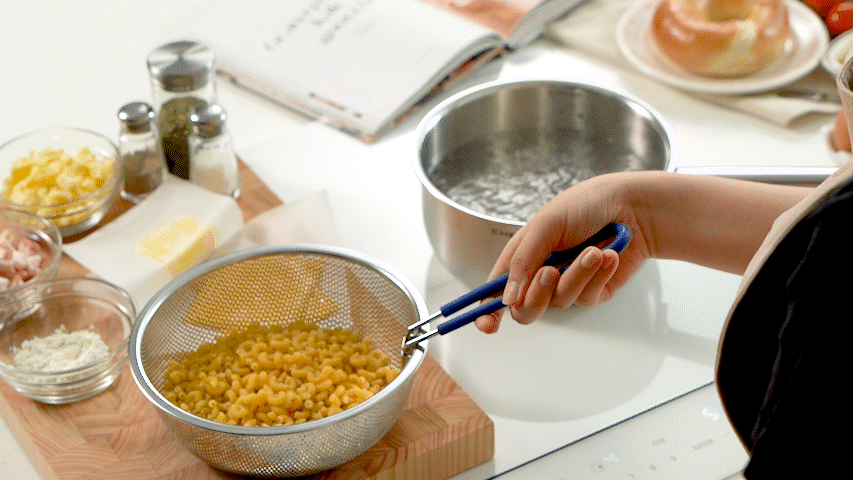 A person drains cooked pasta over a colander in a kitchen, surrounded by ingredients like cheese, spices, and a cookbook.
