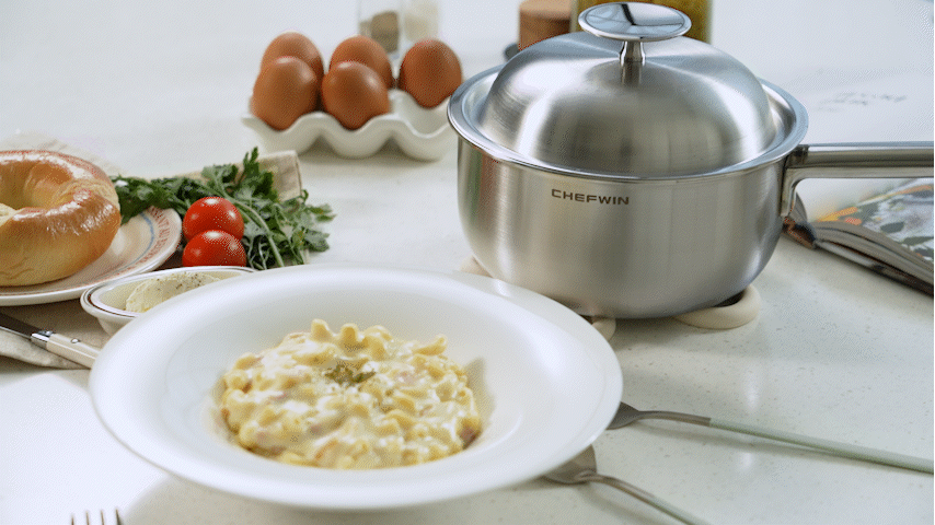 A plate of macaroni and cheese on a kitchen countertop, with a stainless steel pot nearby, eggs in a carton, cherry tomatoes, parsley, and a bagel with cream cheese in the background.