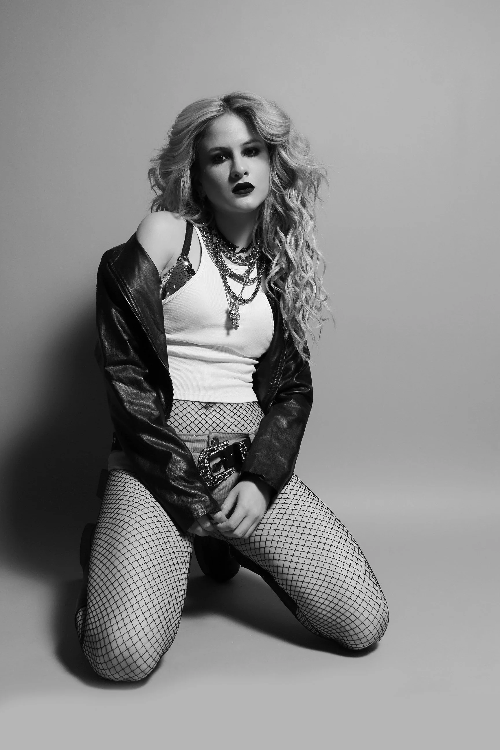 Mujer con cabello rizado largo, maquillaje oscuro, usando una camiseta blanca, chaqueta de cuero, múltiples collares, mallas de red, y sosteniendo una pistola, posando en un fondo gris.
