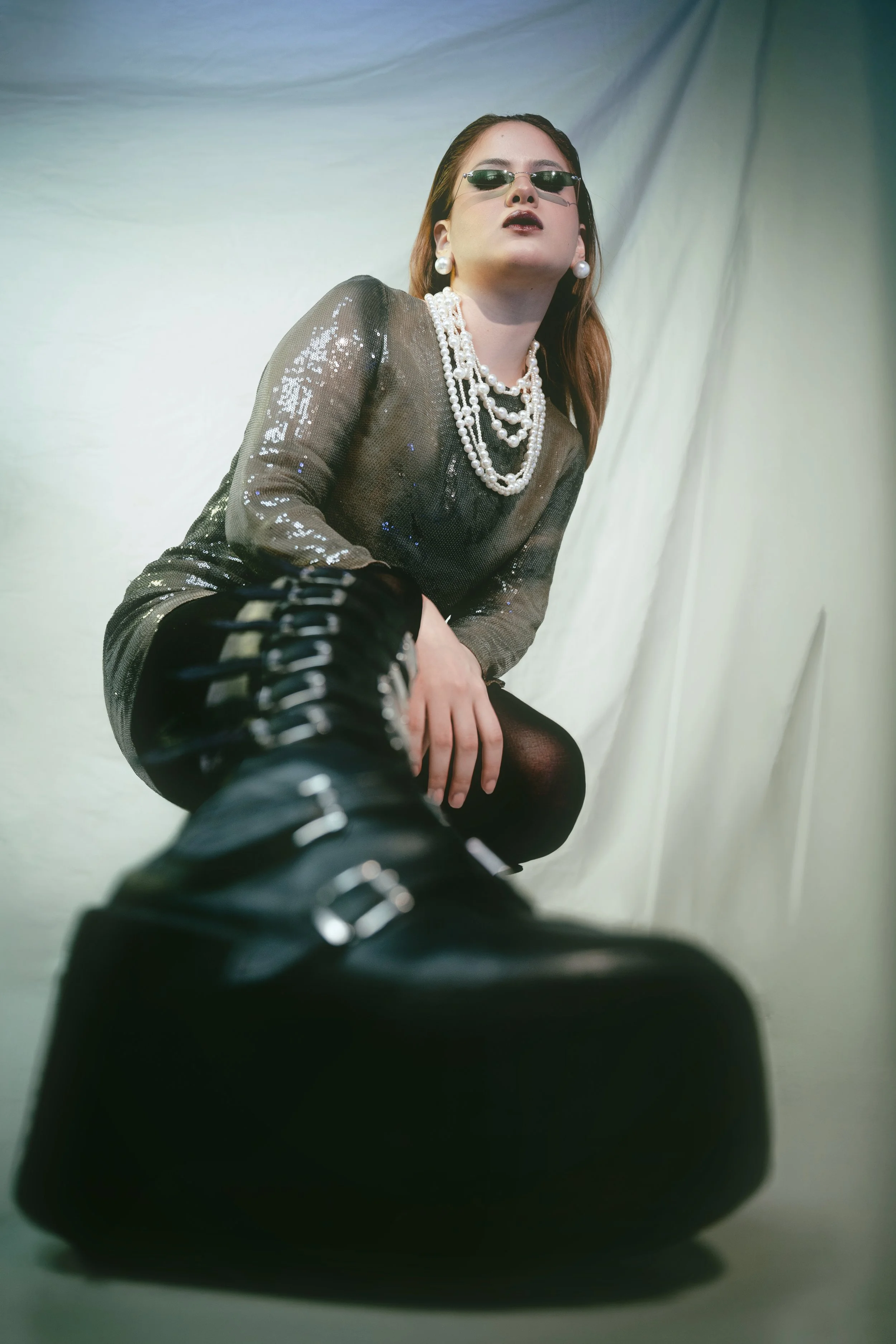 Retrato de una mujer con gafas oscuras, pendientes y múltiples collares, vestida con un vestido brillante y de manga larga, con la espalda apoyada en una pared o cortina de fondo de color gris claro.