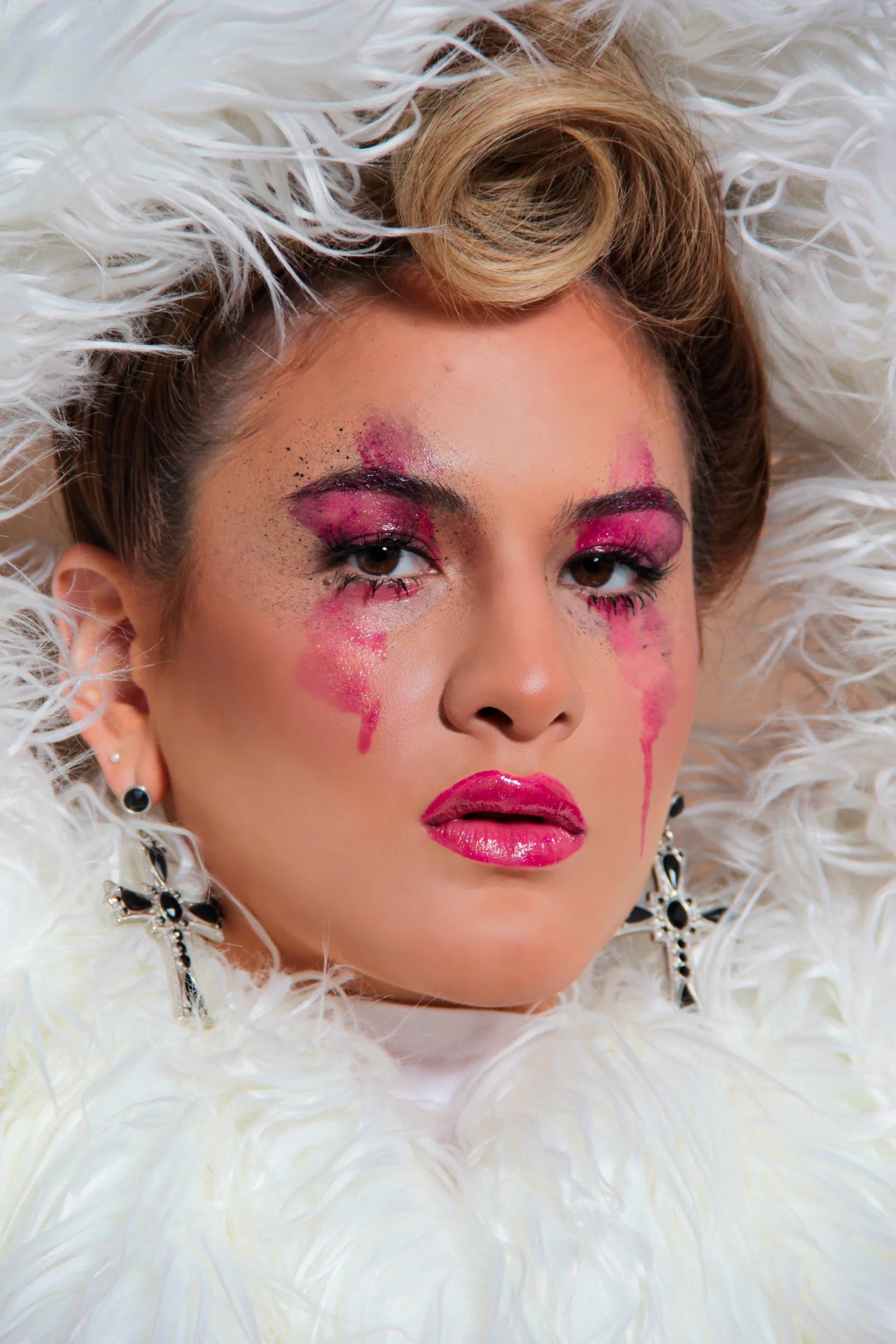 Retrato cercano de una mujer con maquillaje artístico, con lágrimas de pintura rosada en su rostro, rodeada de plumas blancas.