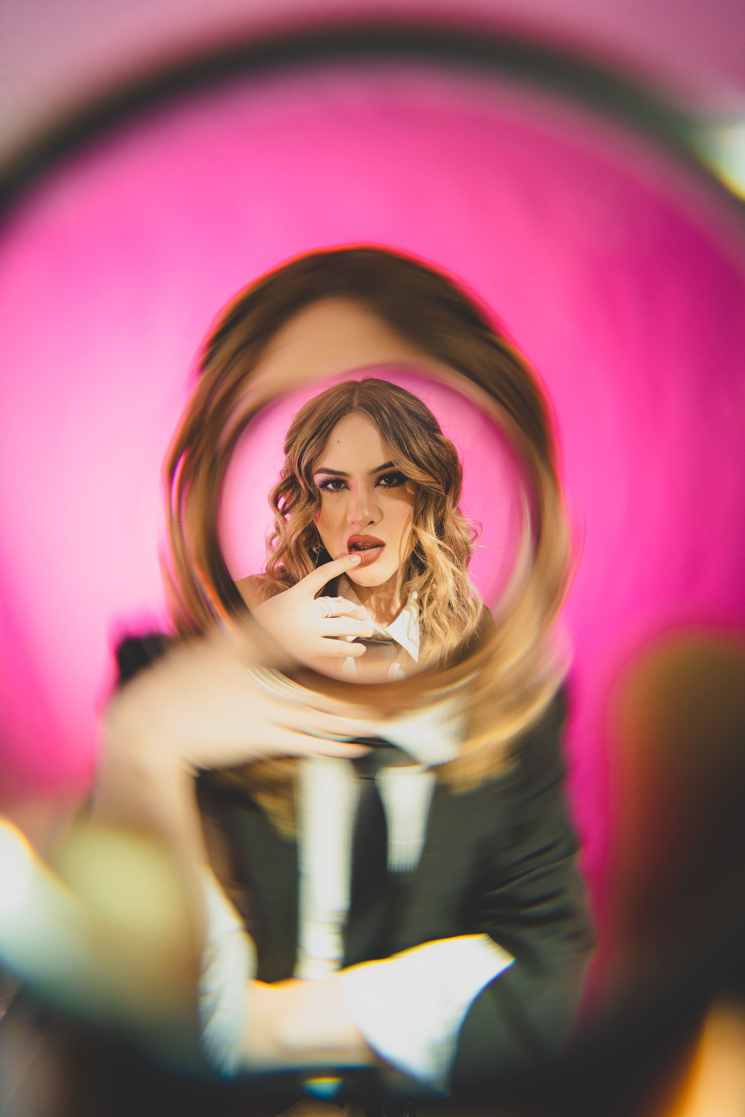 Mujer con cabello castaño y rizado, posando frente a un espejo con marco dorado, tocándose los labios, con fondo rosa.