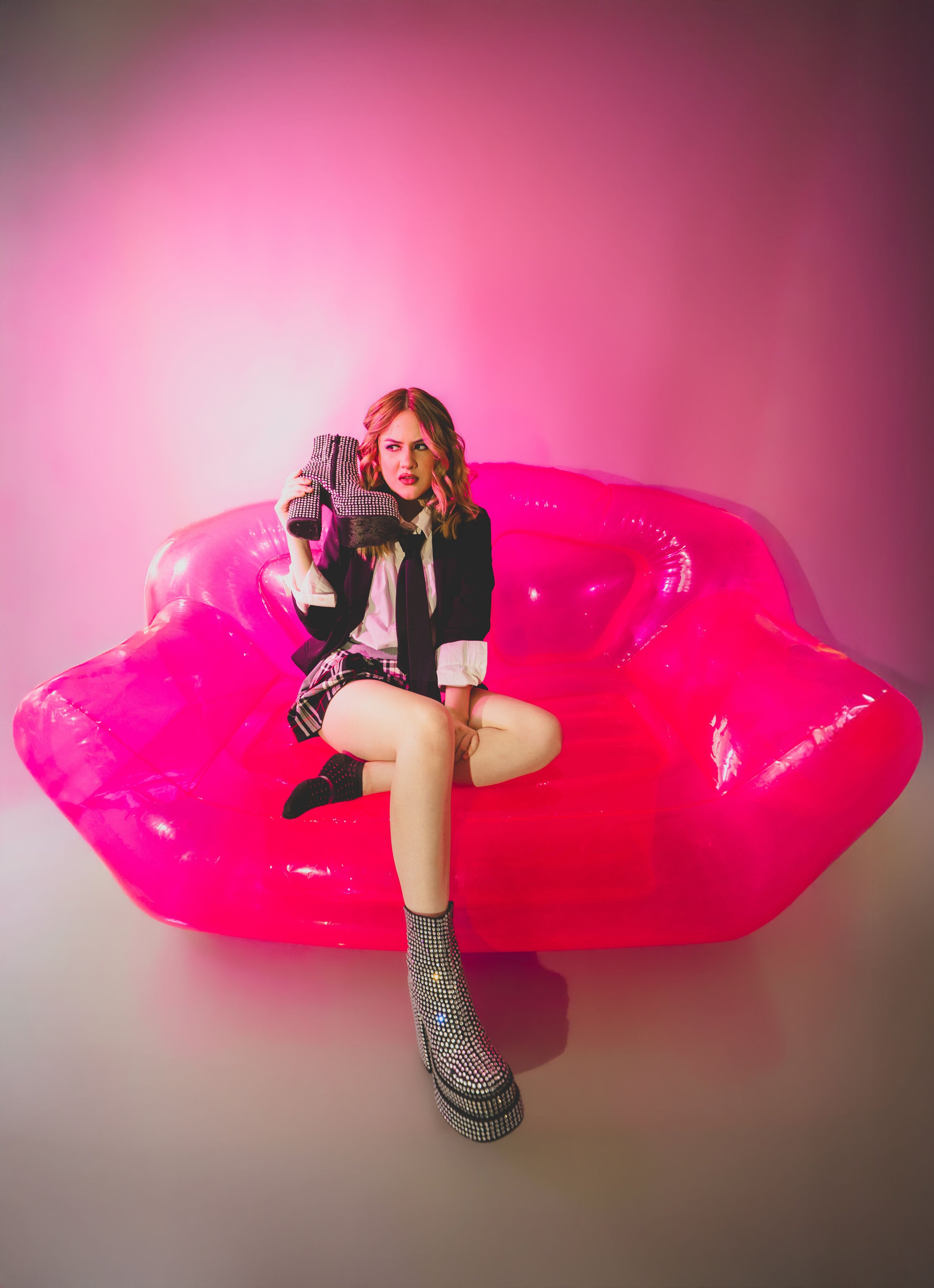 Mujer con cabello rubio, vestida con chaqueta negra y falda a cuadros, sentada sobre un sillón inflable rosa, sosteniendo un zapato de tacón con brillo