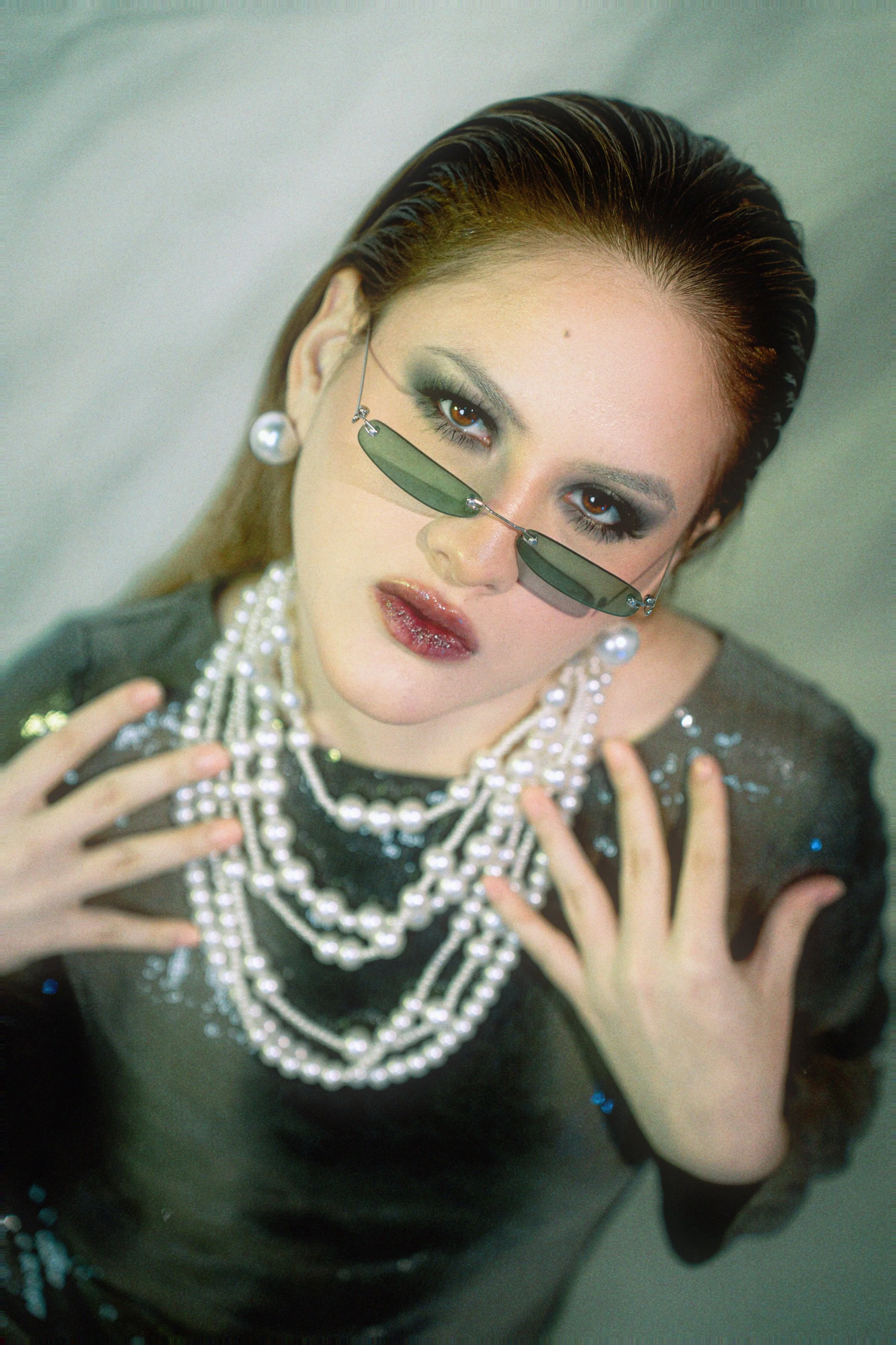 Mujer con peinado mojado, gafas de sol, joyería de perlas y maquillaje oscuro, mostrando el pecho decorado con varias cadenas de perlas.