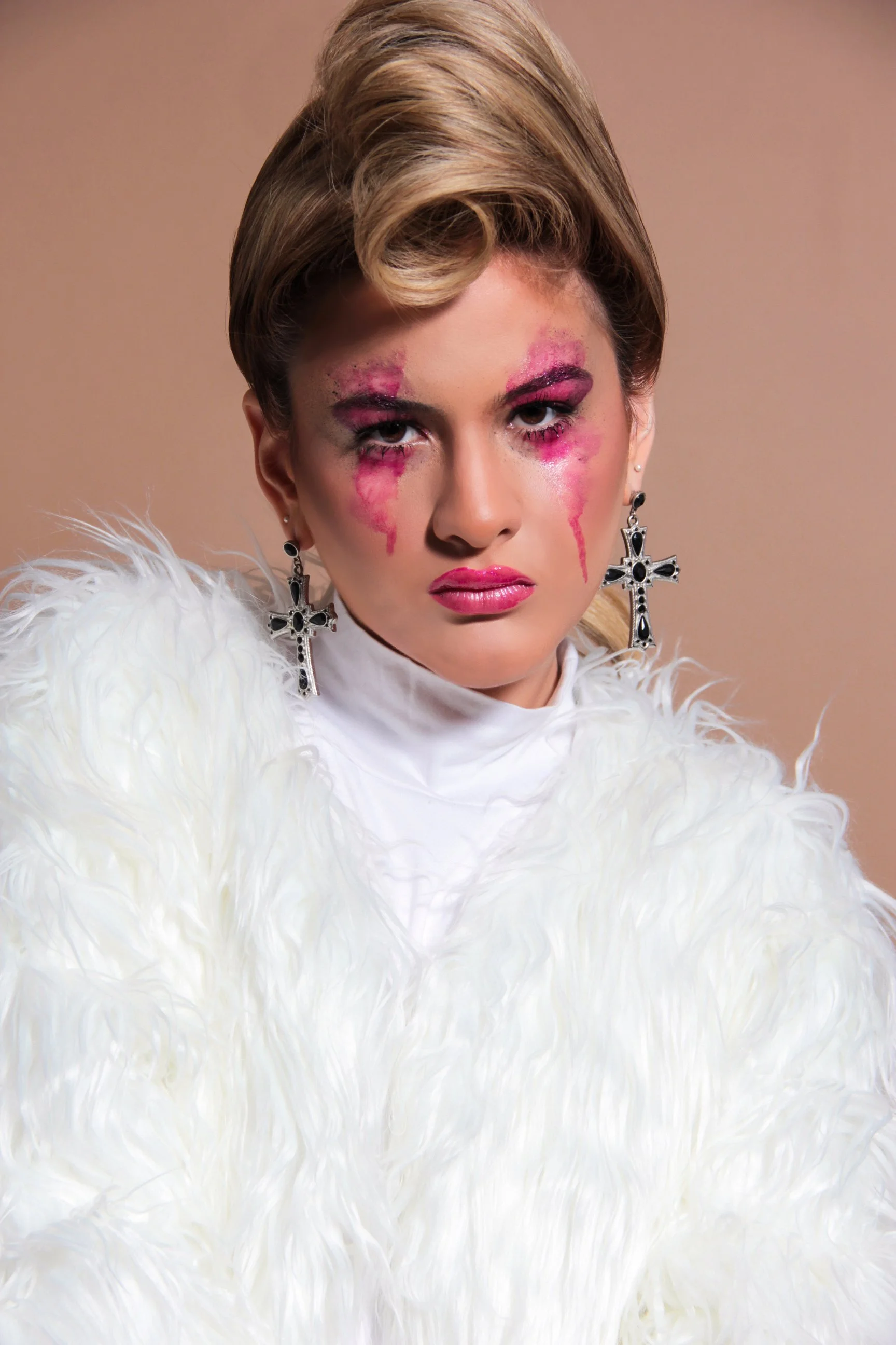 Mujer con maquillaje dramático y lágrimas de pintura rosa, vestida con ropa blanca con plumas largas, usando pendientes de cruz negras y plateadas.