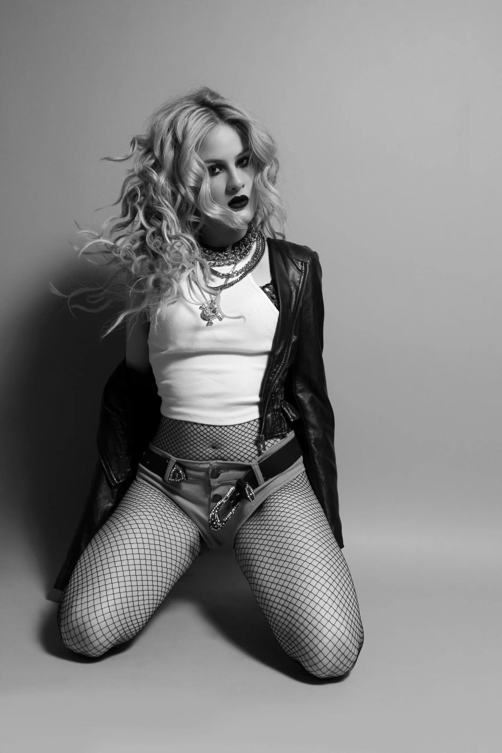 Mujer posando en un estudio, usando ropa provocativa, con cabello rizado, maquillaje oscuro, medias de rejilla y accesorios de joyería, en una pose sensual.