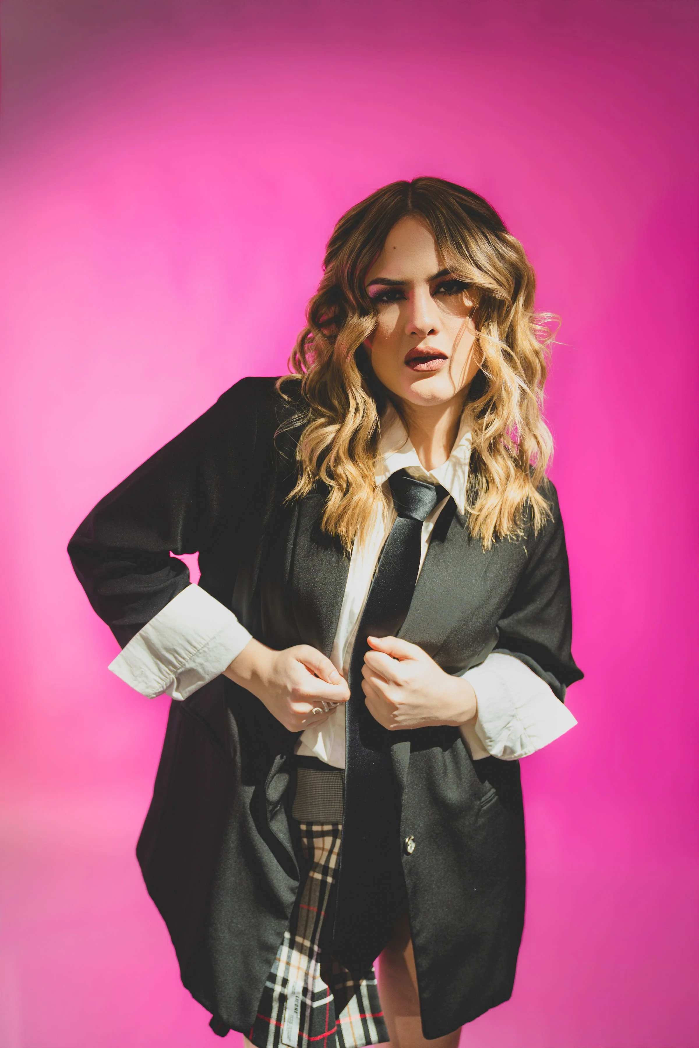 Retrato de una mujer con cabello rubio y ondulado, ropa de estilo formal con blazer negro, camisa blanca y una corbata negra, posando frente a un fondo rosa vibrante.