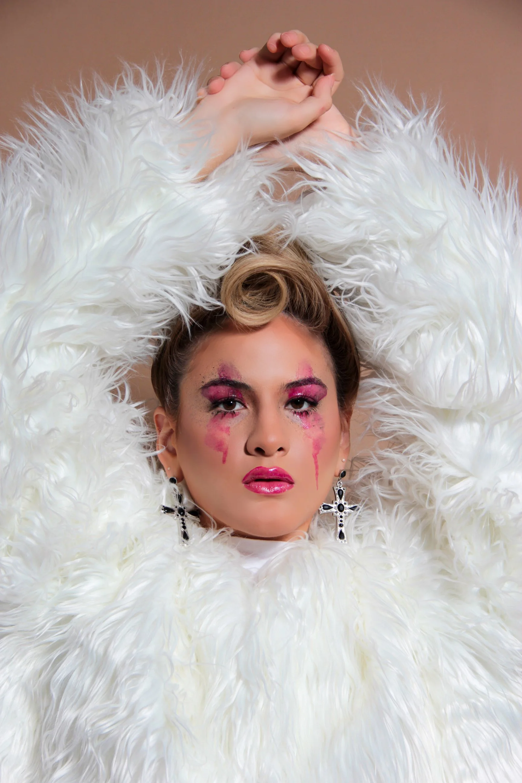 Mujer con maquillaje llamativo y lágrimas de pintura rosa en su rostro, vestida con un conjunto de plumas blancas largas que la rodean y adornos de joyería con cruces.