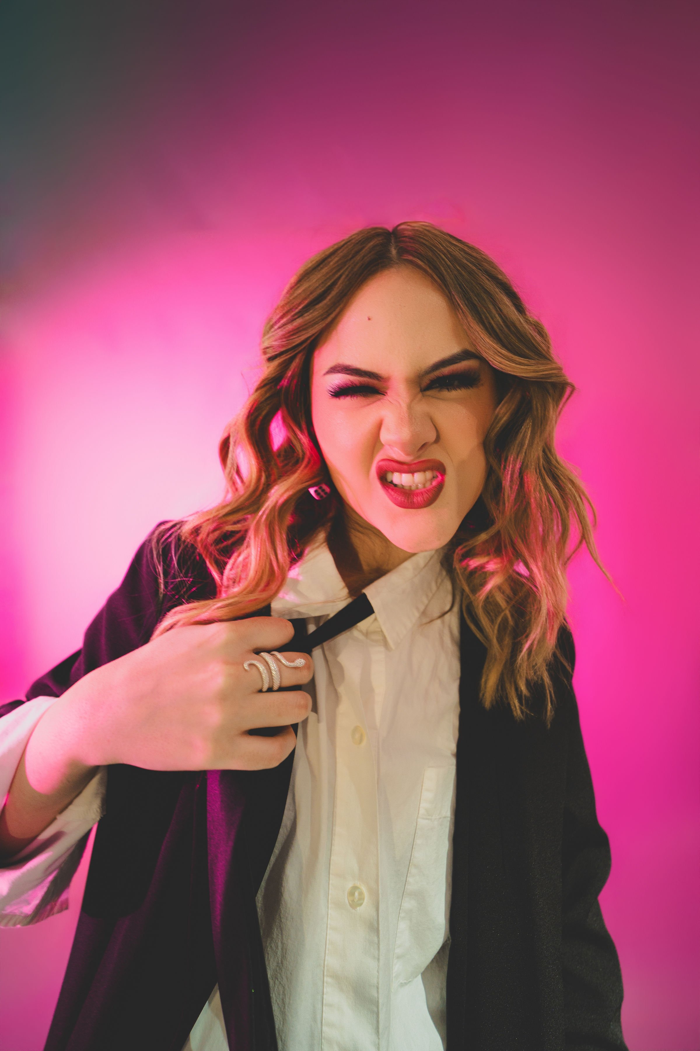 Mujer con cabello rizado, ceño fruncido y expresión desafiante, vestida con camisa blanca y chaqueta negra, con fondo rosa y morado.