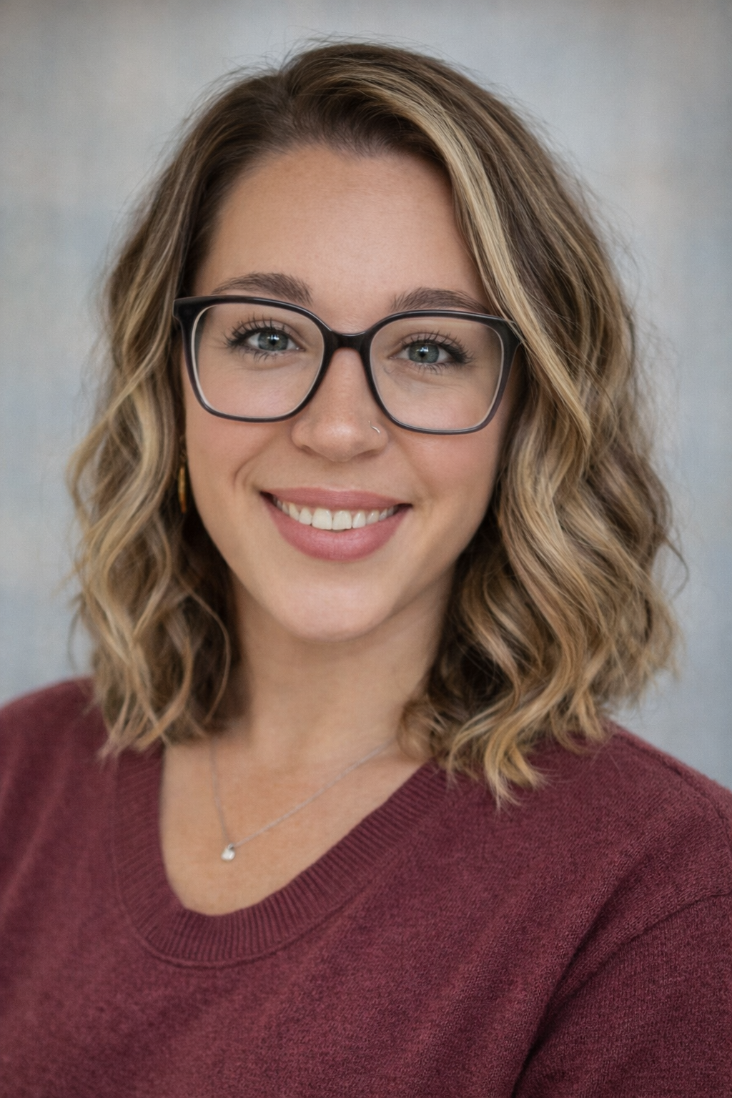 A young woman with wavy, shoulder-length blond hair and glasses, smiling at the camera, wearing a maroon top and a silver necklace.