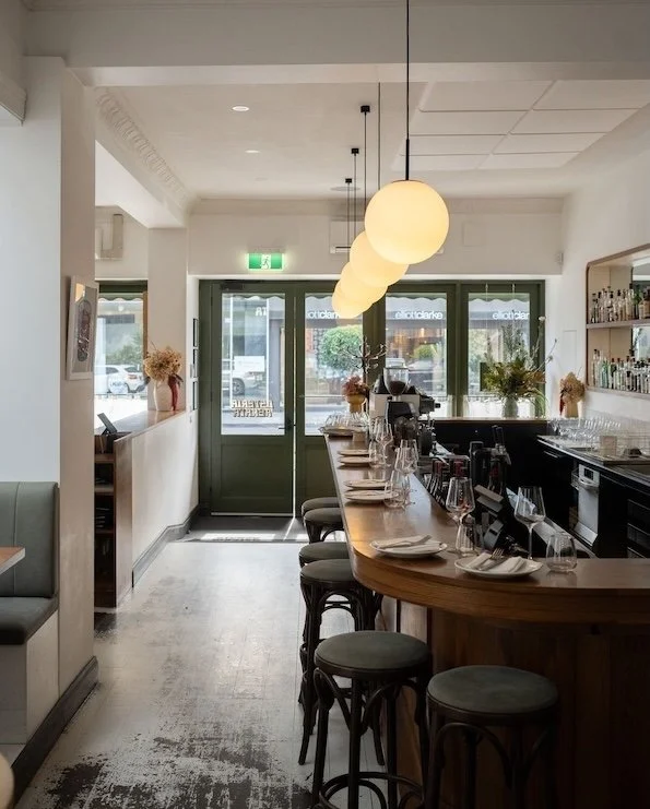 Interior of a cozy restaurant with a curved bar counter, hanging round pendant lights, and decorative plants near windows. Door with glass panels and sidewalk outside visible. Italian flair.