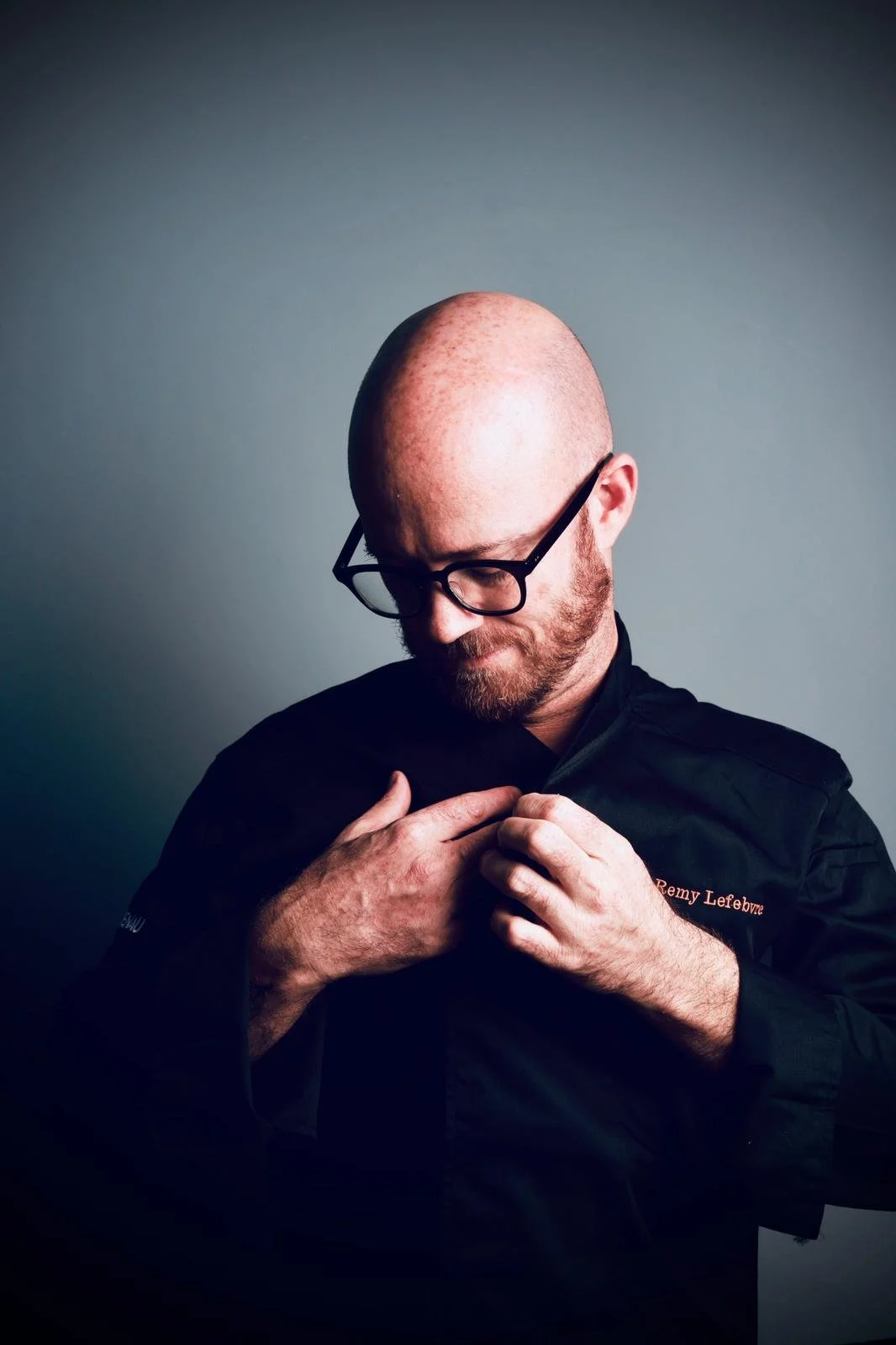 A man with a bald head, glasses, and a beard adjusting his chef's coat that has 'Remy Lefebvre' embroidered on it, standing against a plain gray background.